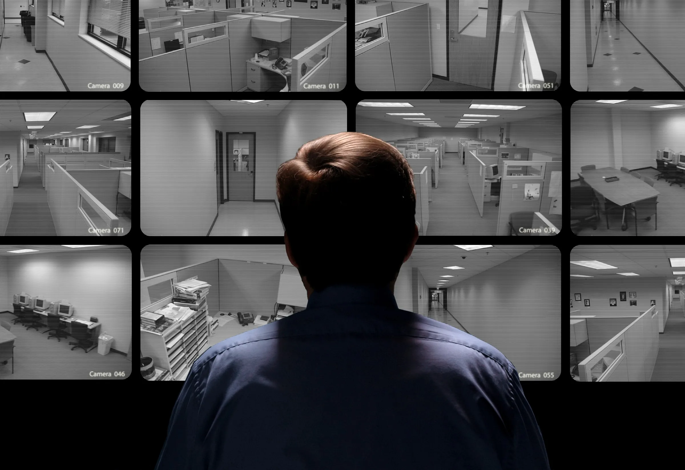 A security officer in a dark uniform stands with arms crossed, watching a wall of monitor screens showing store camera feeds