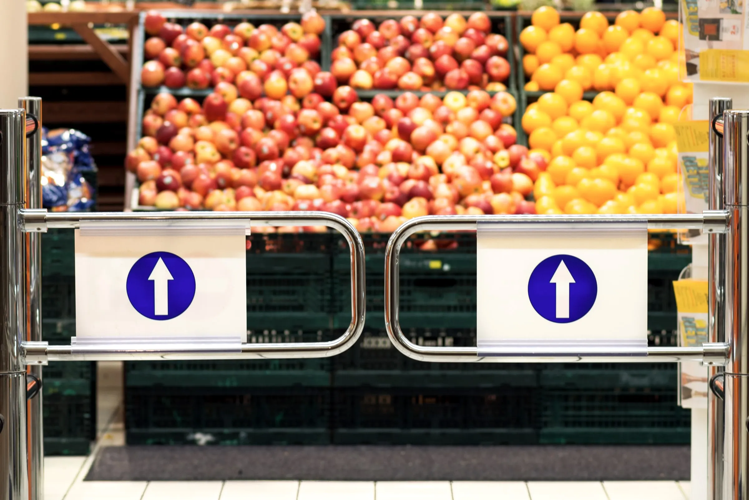 A chrome one-way entry gate with blue directional arrows stands in front of a blurred grocery produce display of apples and oranges