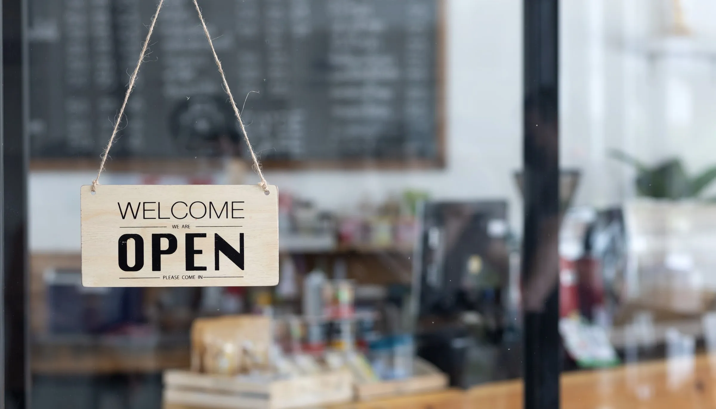 Wooden welcome sign reading we are open hanging by twine on a glass shop door, with a blurred counter, register and chalkboard behind