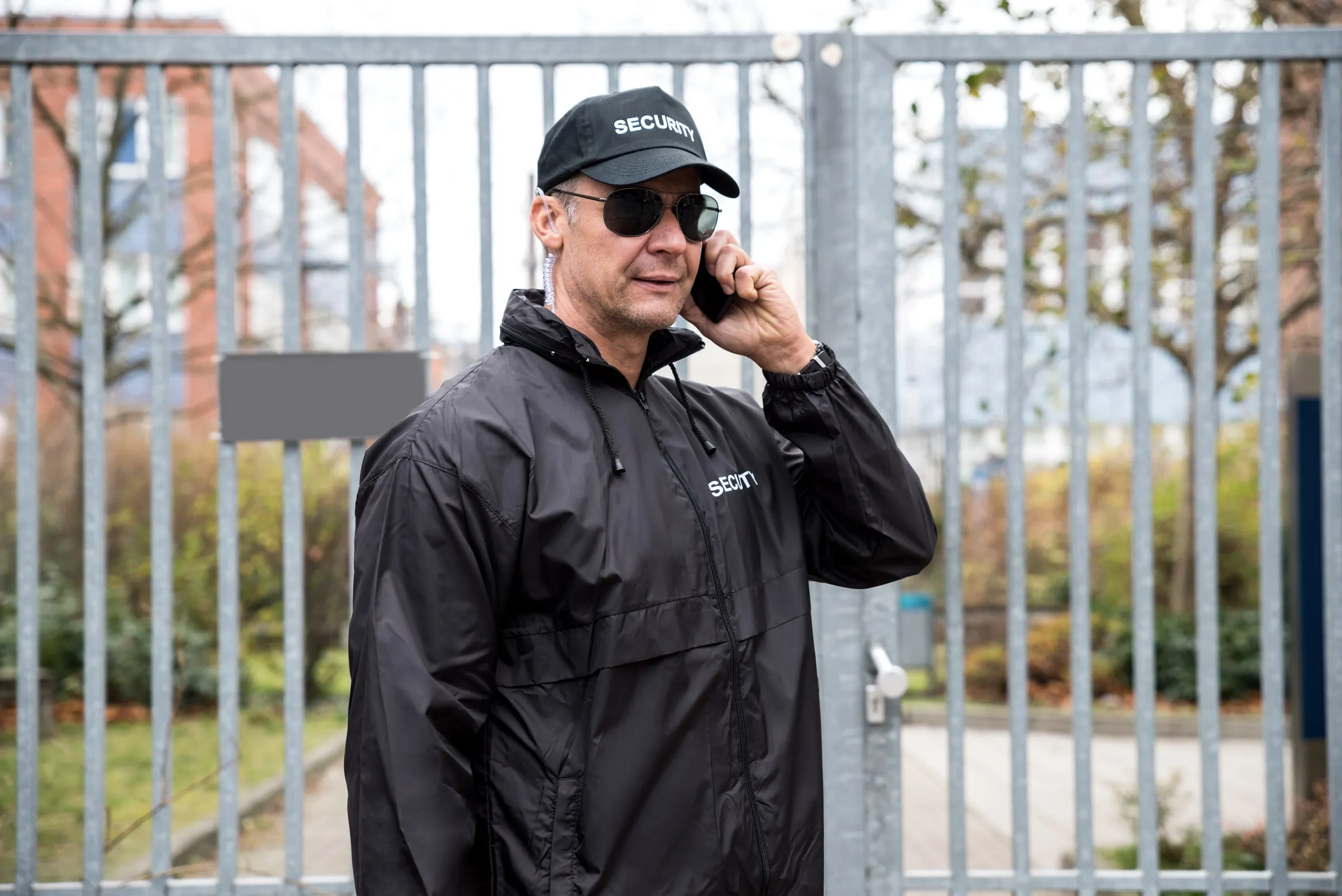 A uniformed security officer in a cap marked security and sunglasses talks on a phone at a metal gate