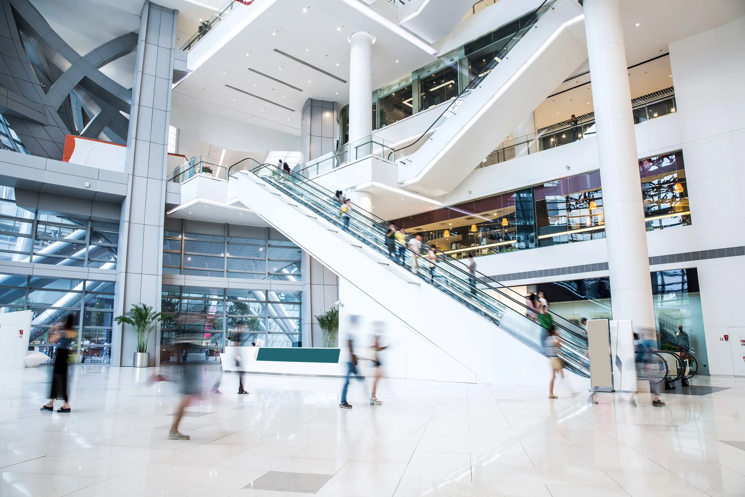 Multi-level shopping mall atrium with a long escalator, white columns, glass railings and blurred shoppers crossing a polished floor