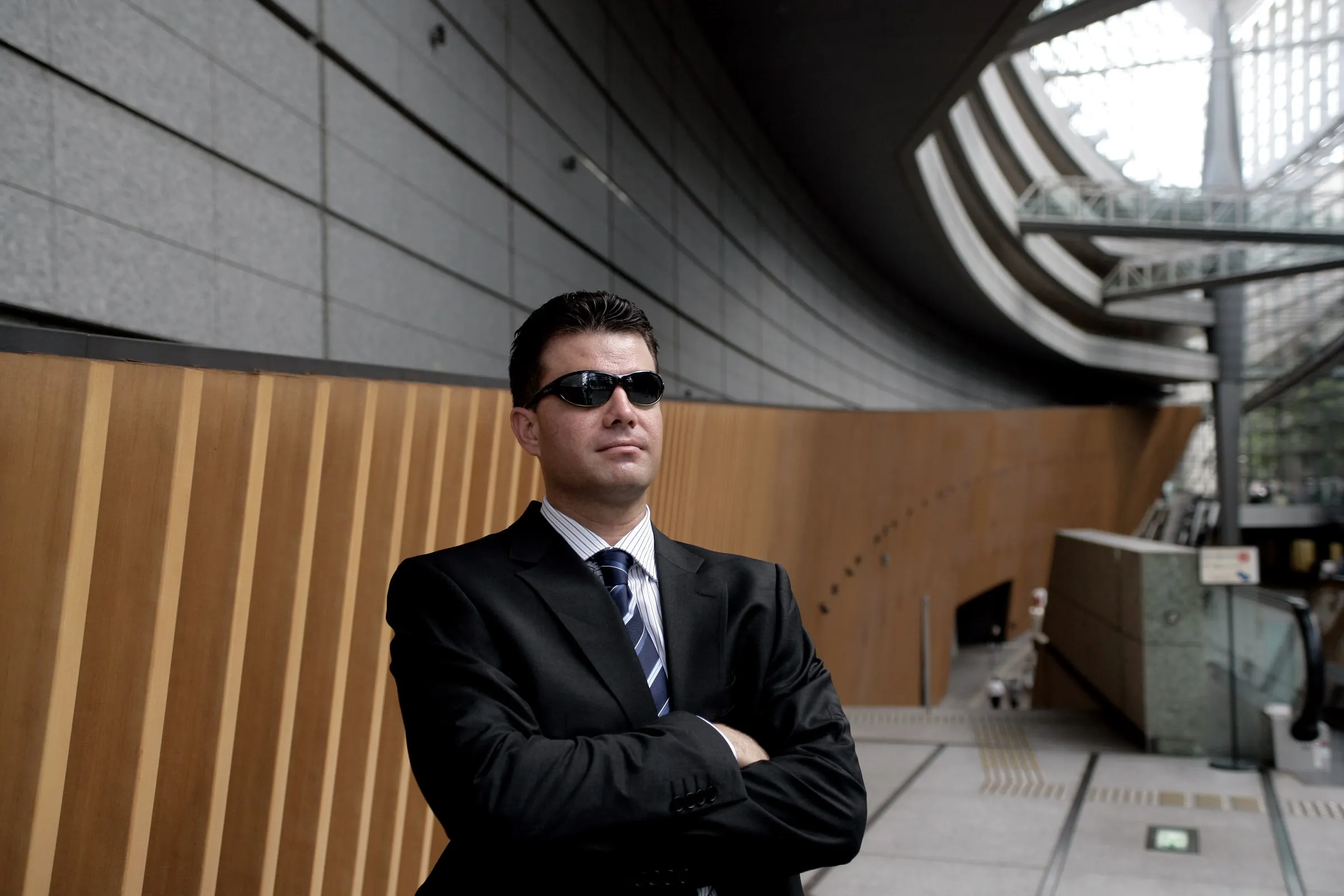 Man in a dark suit and sunglasses standing with arms crossed in a glass-roofed atrium with a curved wooden wall