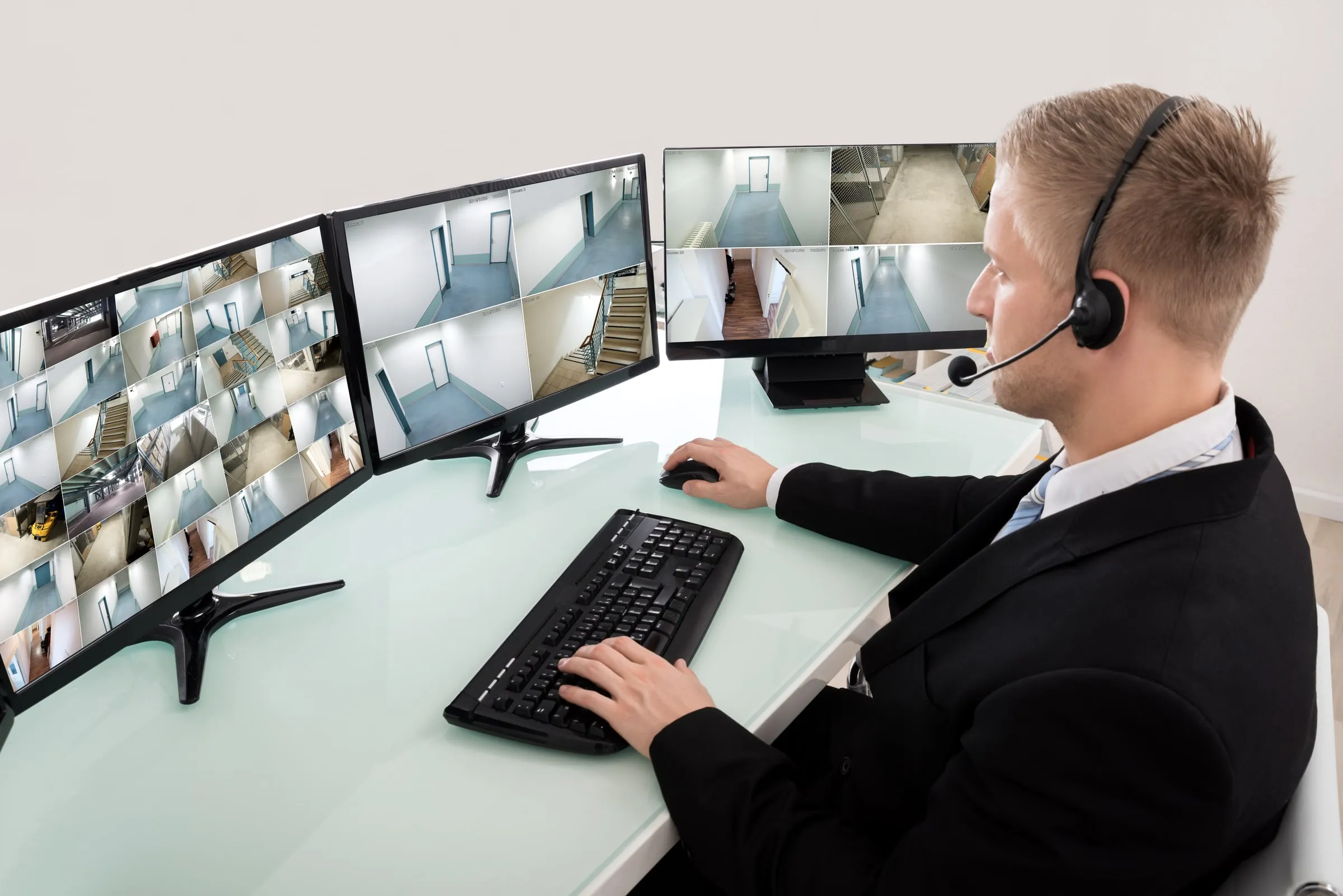 A man in a suit sits at a desk watching a wall of security camera monitors showing building interiors, wearing a headset