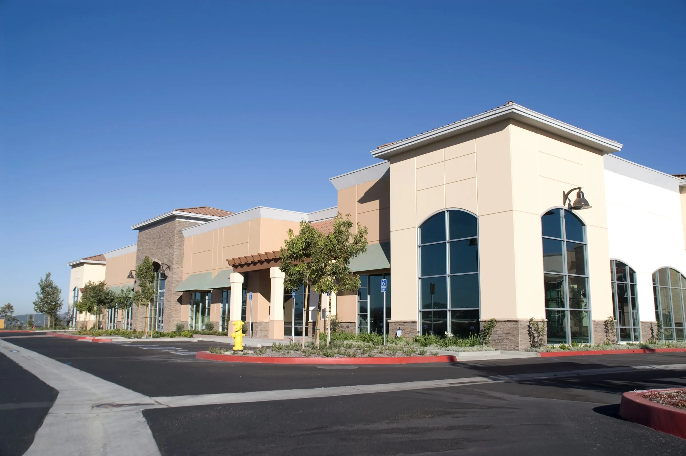 Single-storey strip retail center with tan stucco, arched windows and green awnings beside an empty parking lot under a clear blue sky