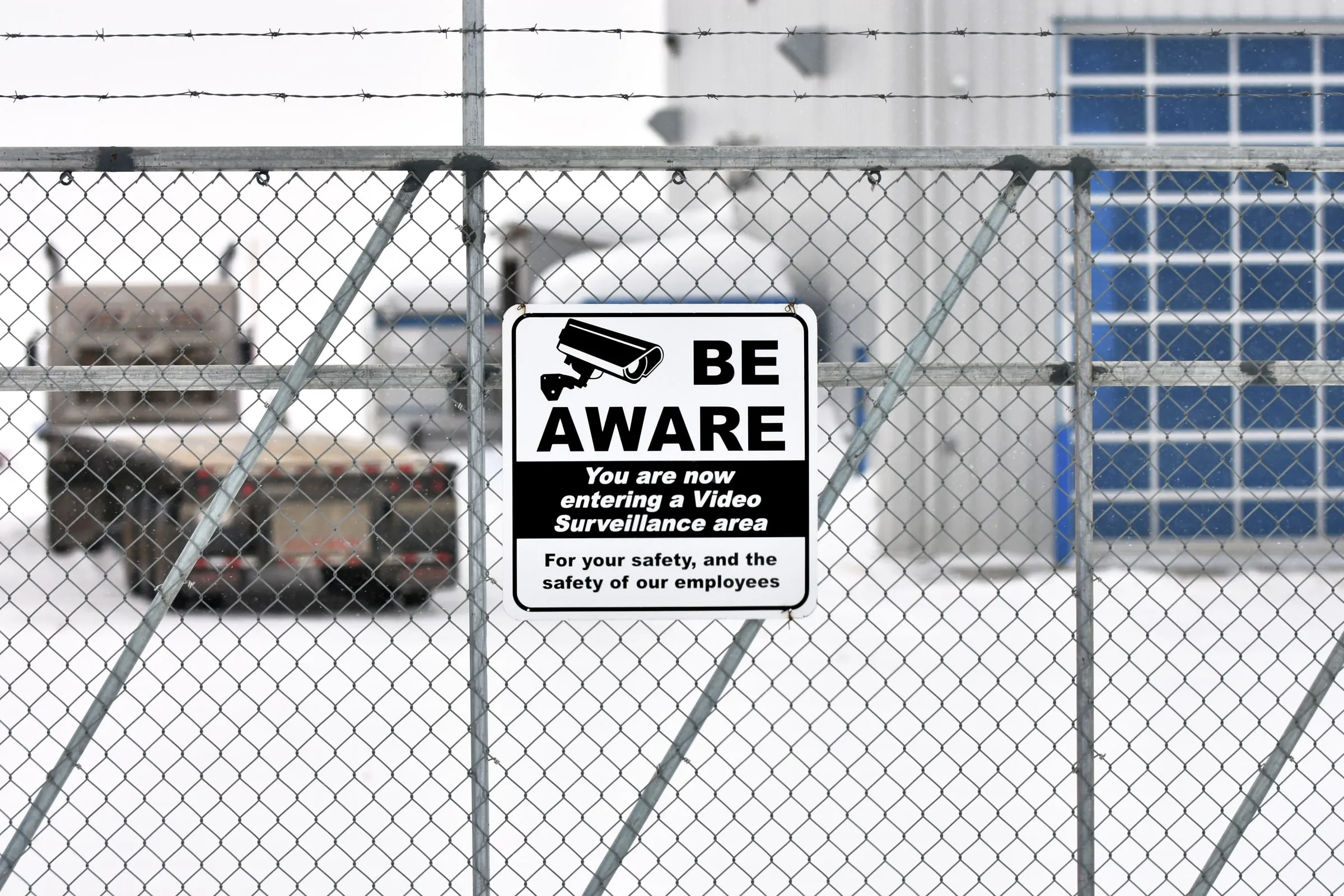 A be aware video surveillance sign hangs on a chain-link fence at an industrial yard with a parked truck