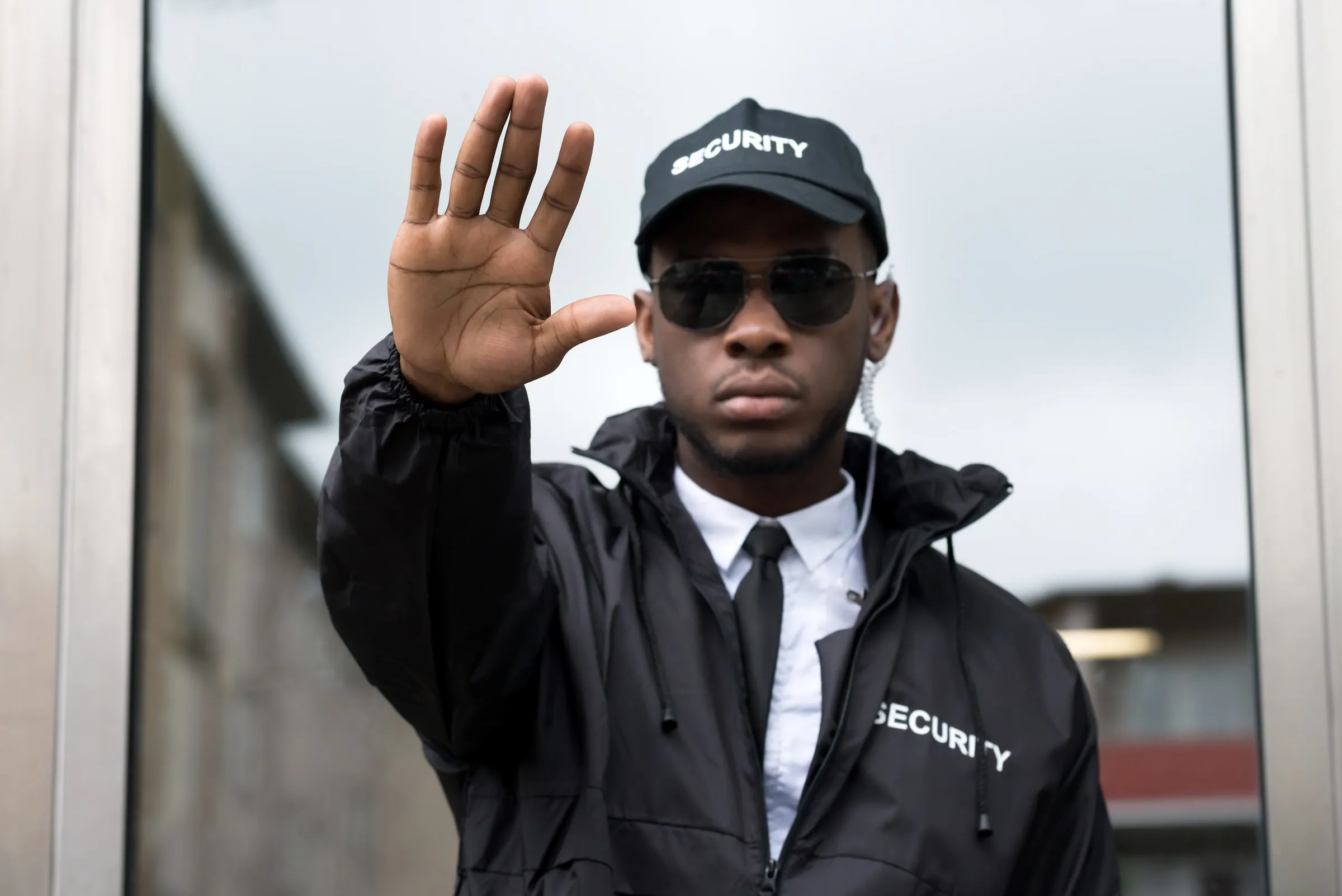 Security officer in a black SECURITY jacket and cap holding up an open palm in a stop gesture at a doorway