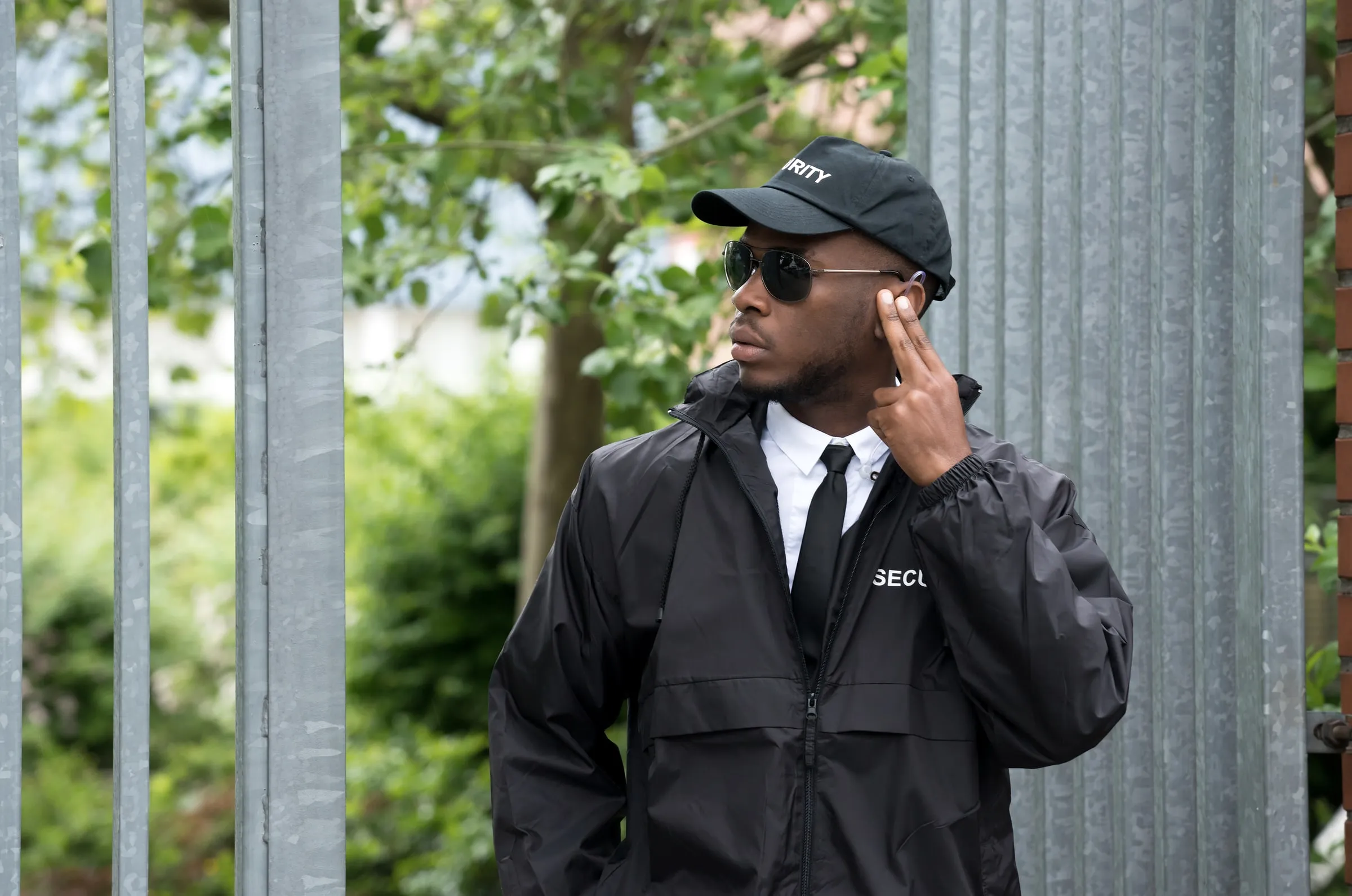 Security officer in a black jacket and cap touching an earpiece beside a metal fence