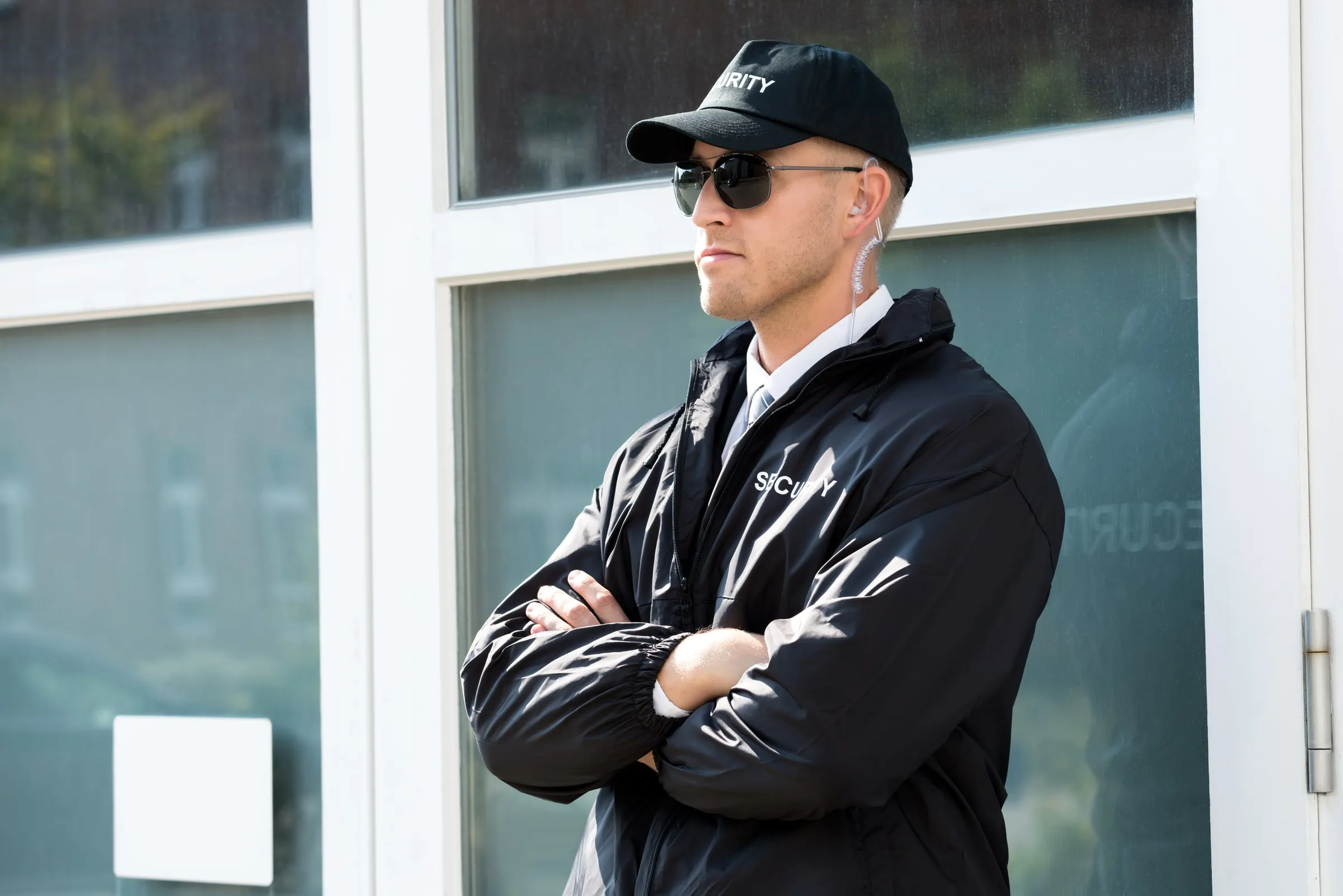 Security officer in a black jacket and cap with arms crossed, standing at a glass office entrance