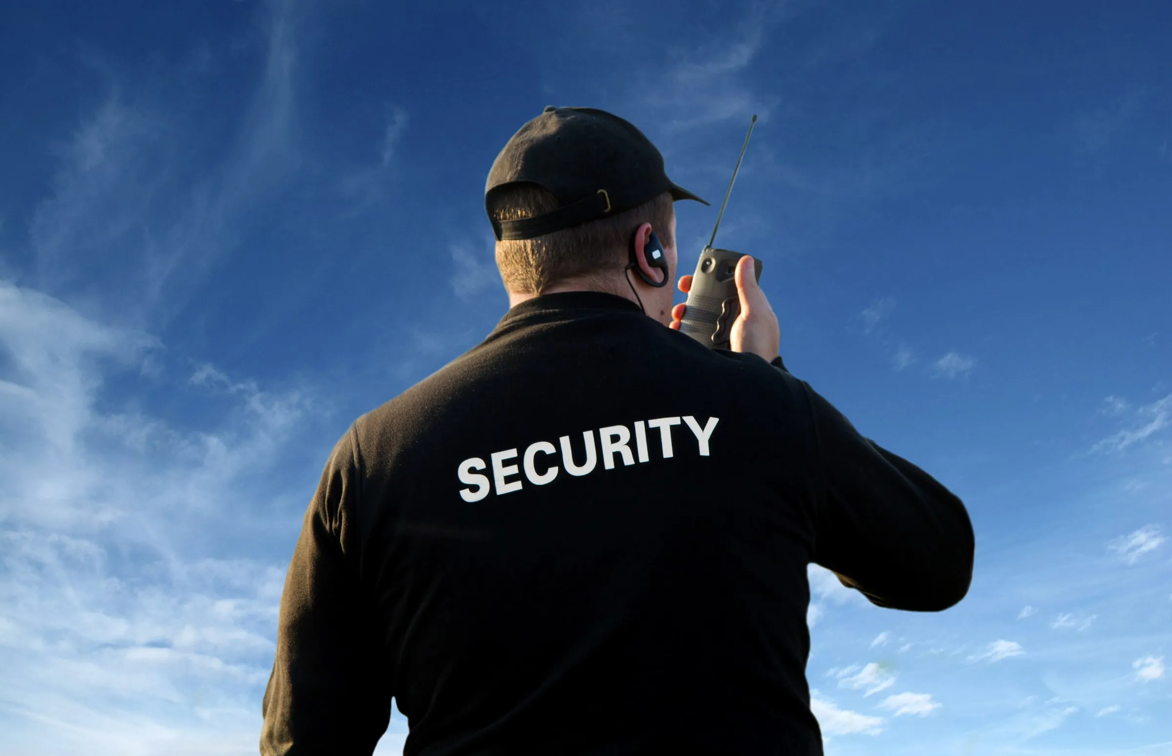 Security officer in a black SECURITY shirt speaking into a radio against a blue sky