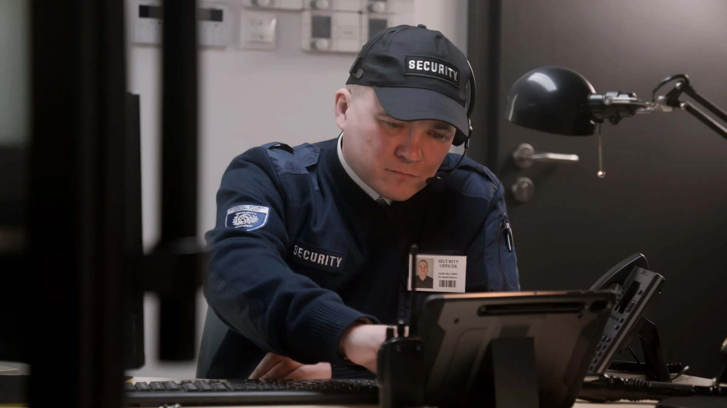 Security officer in a dark uniform at a desk under a lamp, writing on a tablet