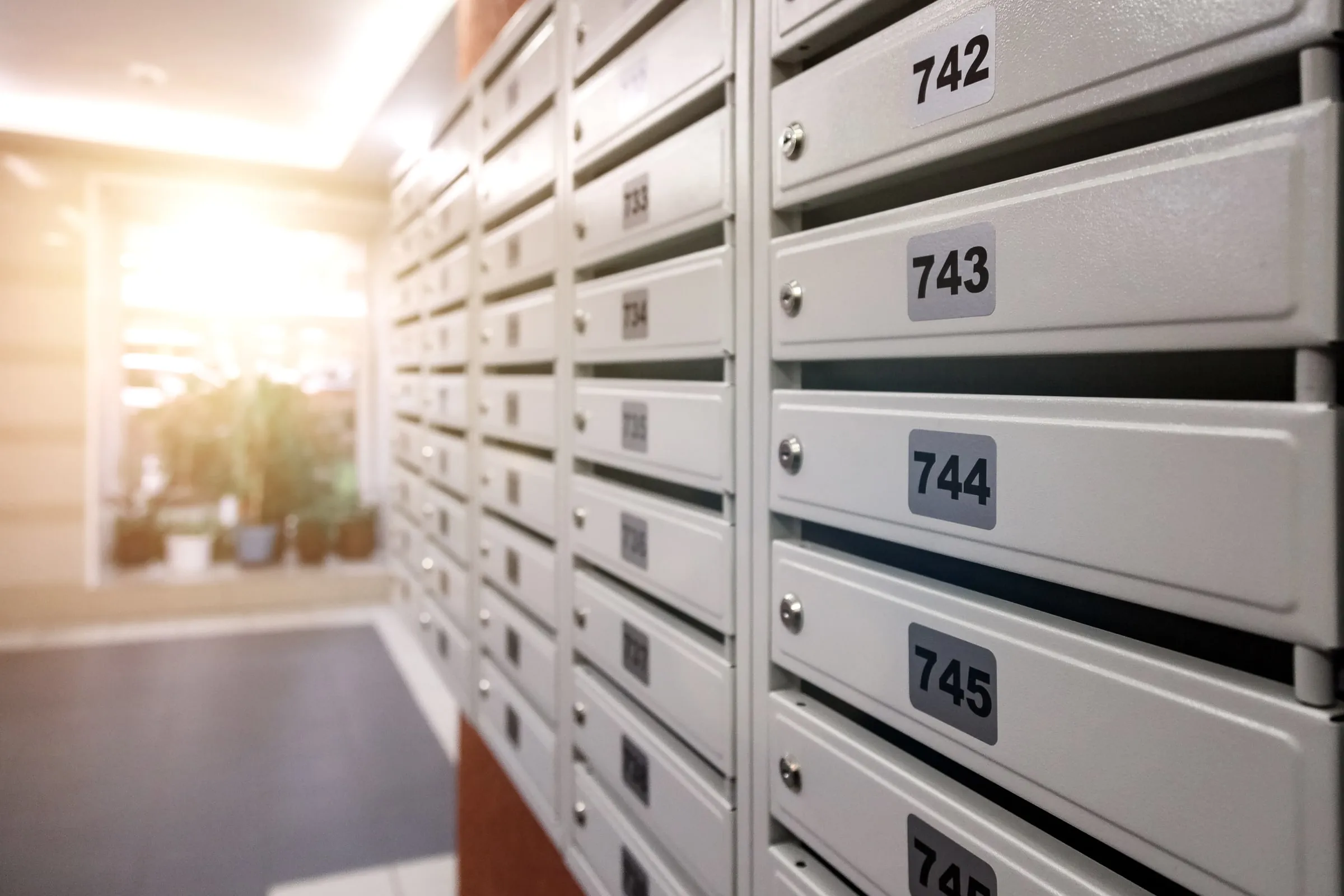 Wall of numbered resident mailboxes in an apartment building's sunlit entry breezeway, with potted plants visible near the glass doors beyond