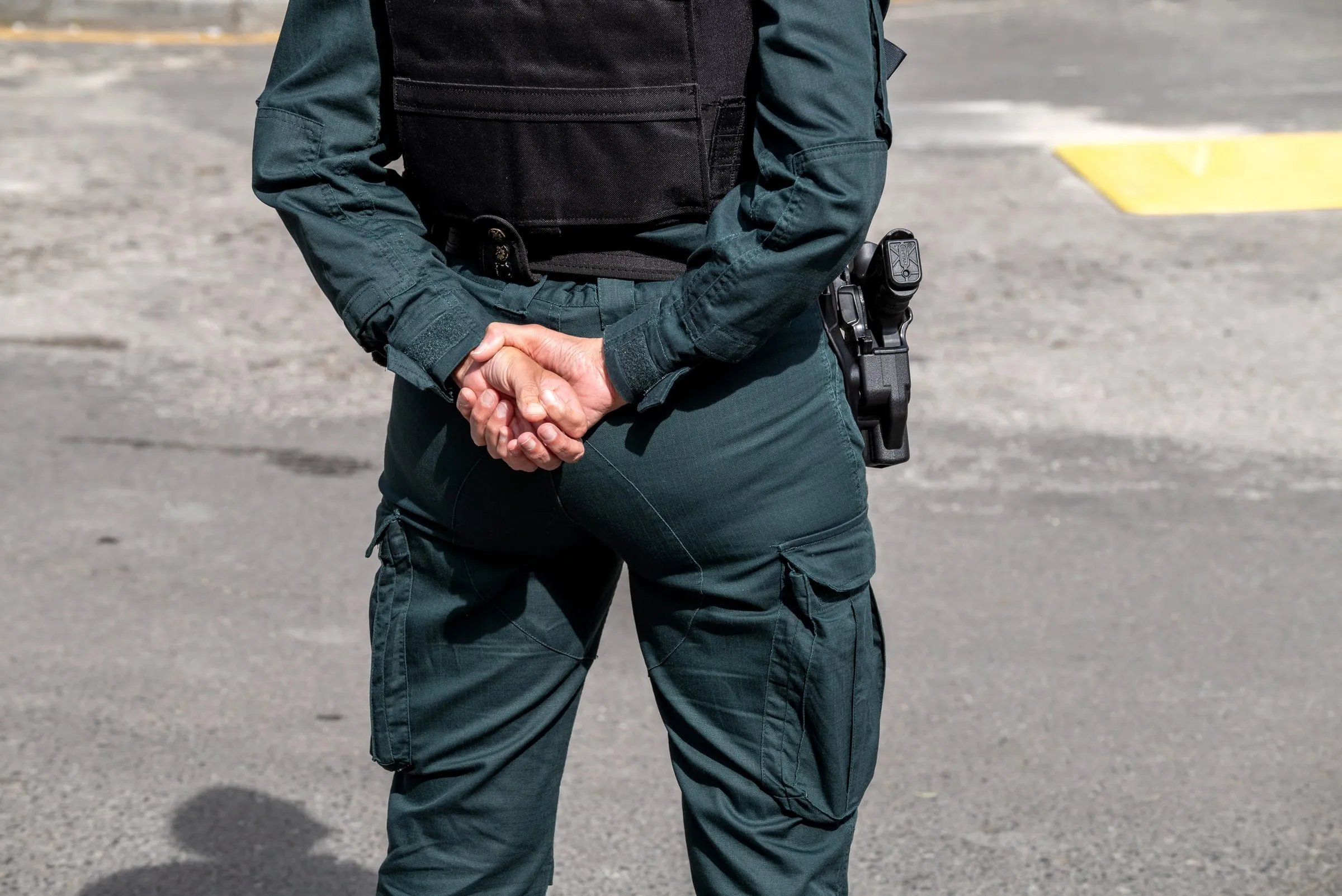 Close view of a uniformed officer's holstered handgun and hands clasped behind the back in a parking lot
