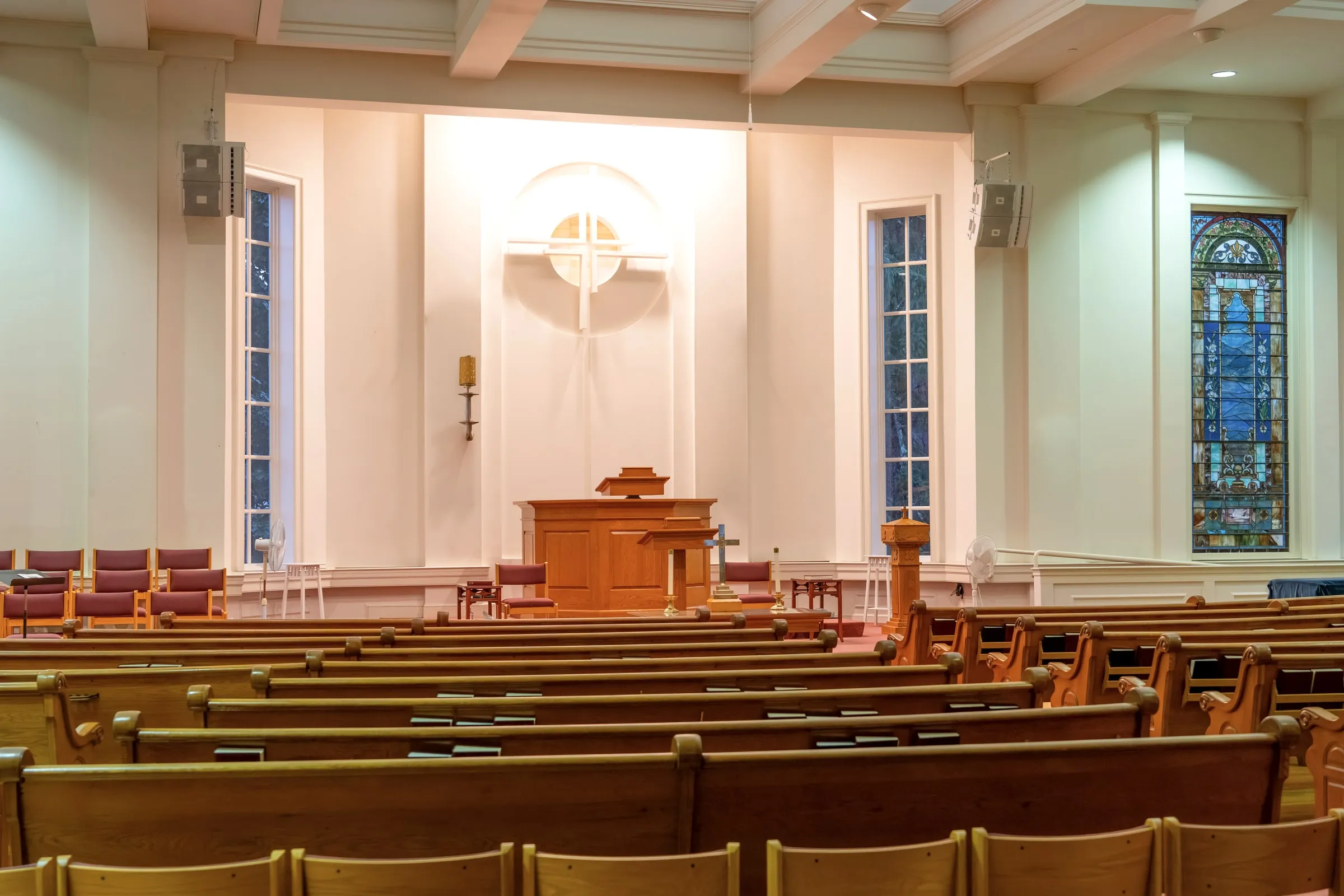 Empty church sanctuary with wooden pews facing a pulpit beneath a circular cross-shaped window and a stained glass window along the side wall