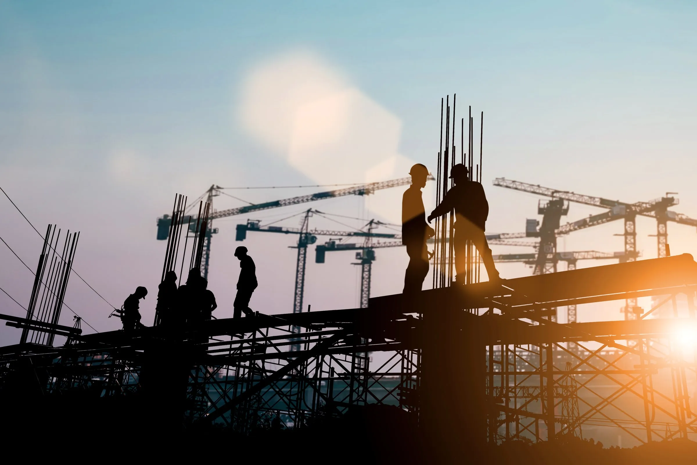 Silhouetted construction workers in hard hats on an elevated steel beam with tower cranes against a hazy evening sky