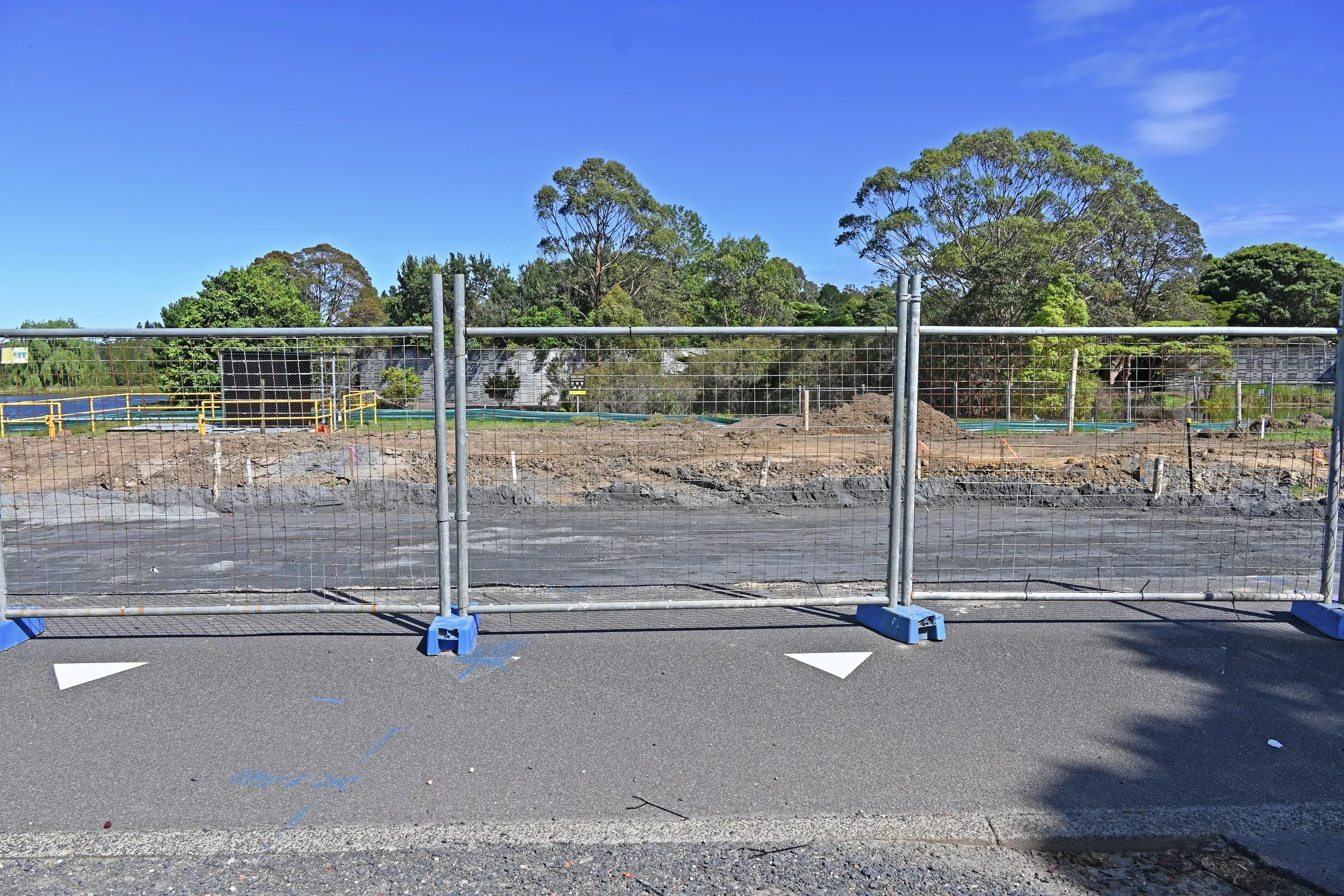 Temporary steel-mesh construction fencing along a cleared building site, with a partly built pool structure and mature trees visible beyond under a clear sky