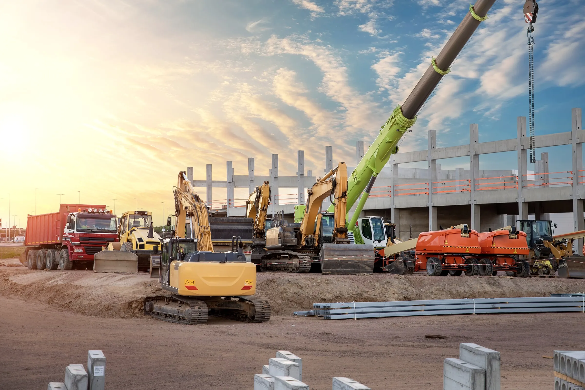 Excavators, a dump truck, a roller and a mobile crane lined up at a construction site at dusk, with a precast concrete parking structure behind them