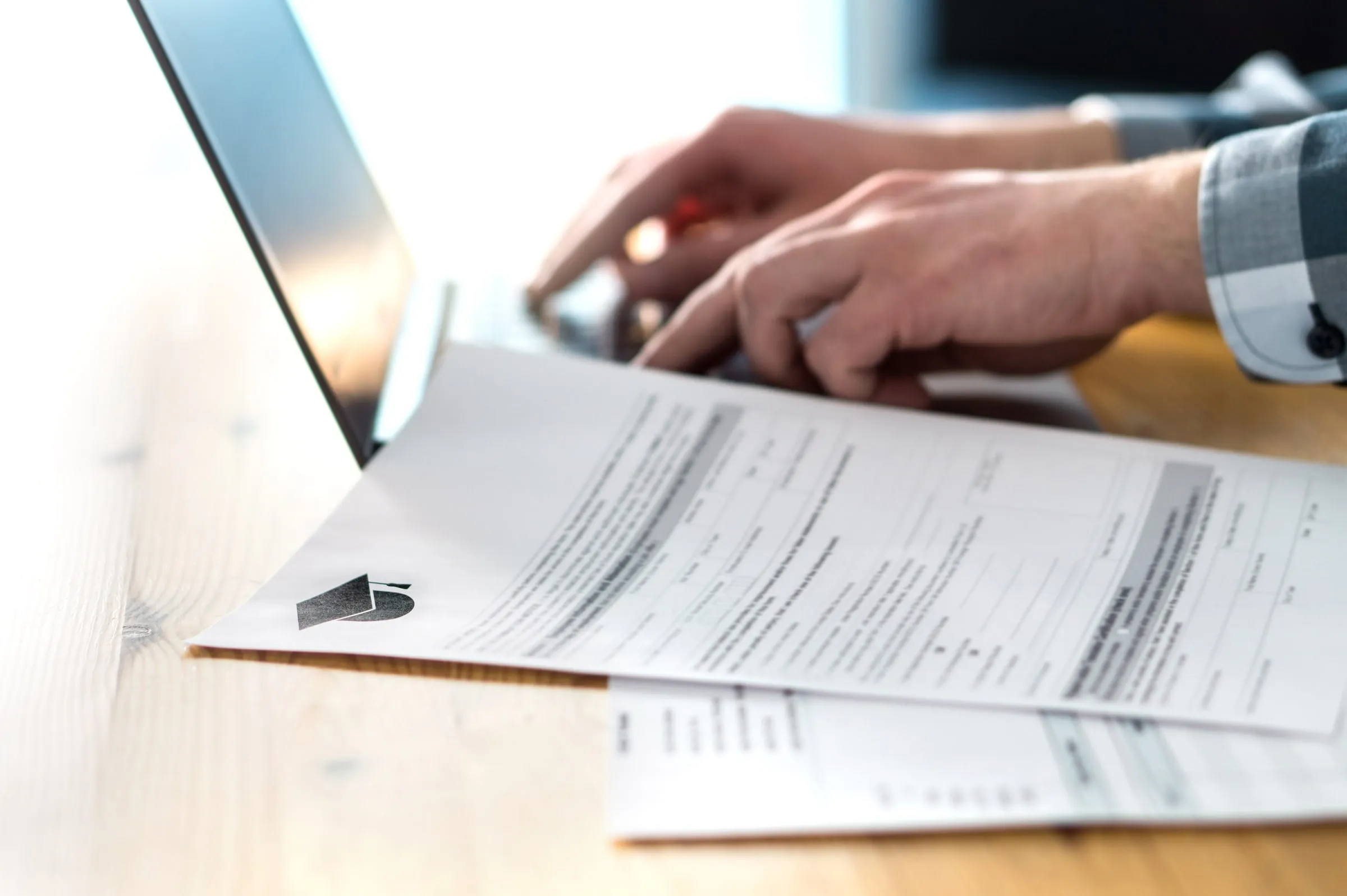 Close view of hands typing on a laptop beside a printed form on a wooden desk
