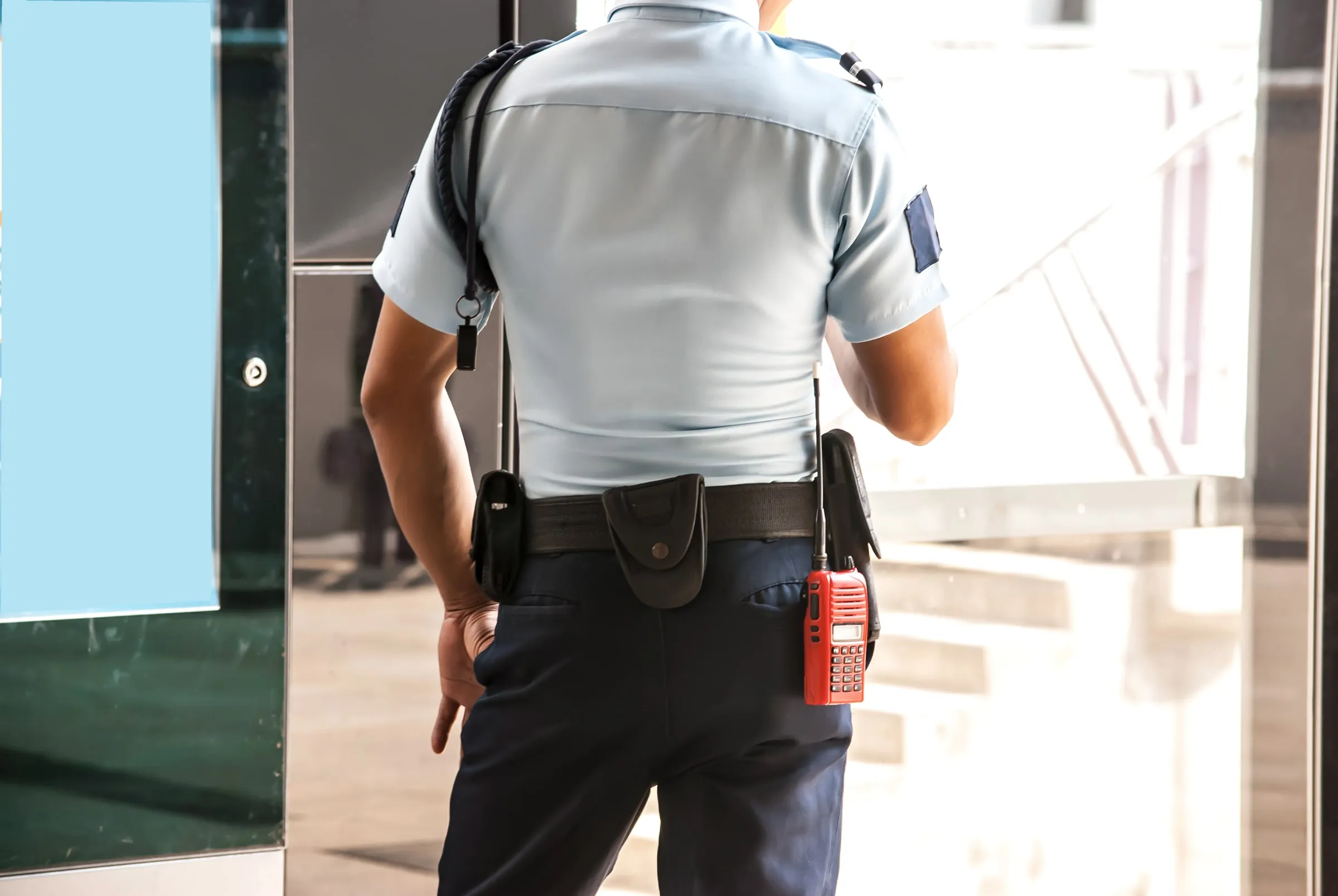Rear view of a uniformed officer standing with hands on a duty belt holding a radio, facing toward a bright building entrance