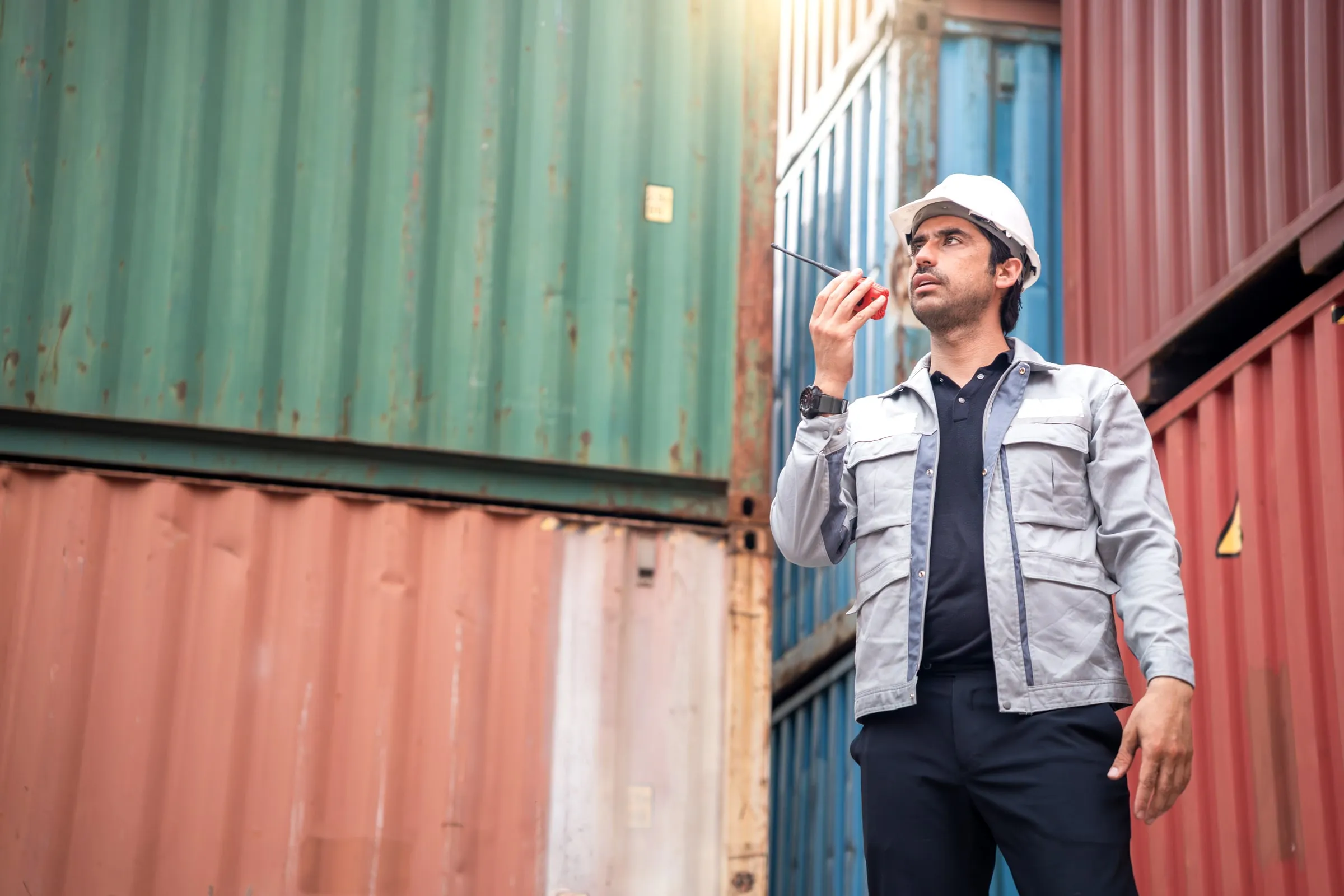Worker in a hard hat and jacket speaking into a handheld two-way radio between rows of stacked shipping containers