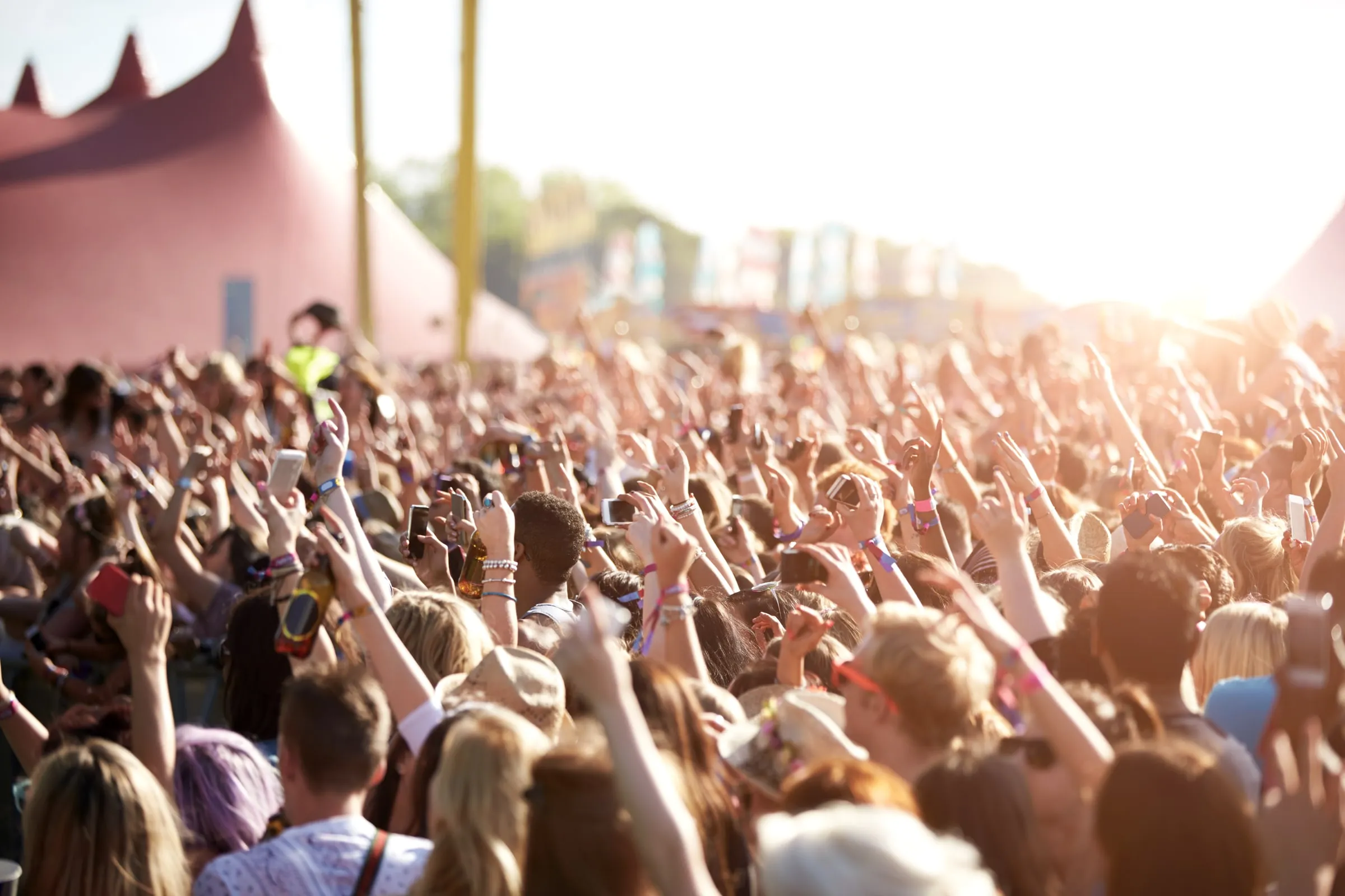Dense outdoor festival crowd with arms raised and phones out, a staff member in a high-visibility vest visible near the stage tents