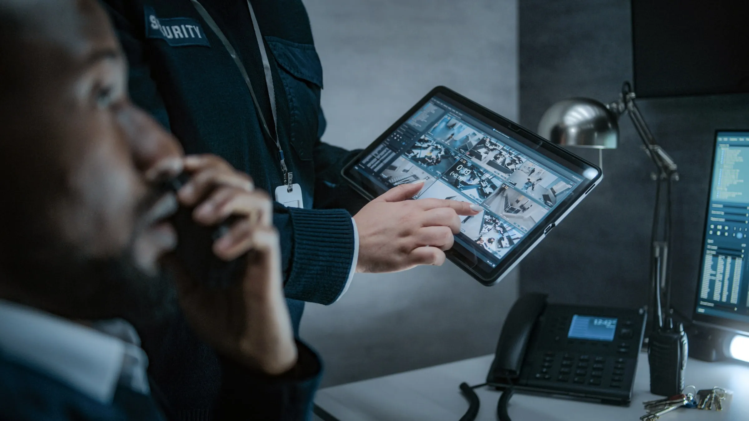 Security officer in a dark jacket pointing to a tablet showing multiple camera feeds while a colleague holds a radio nearby