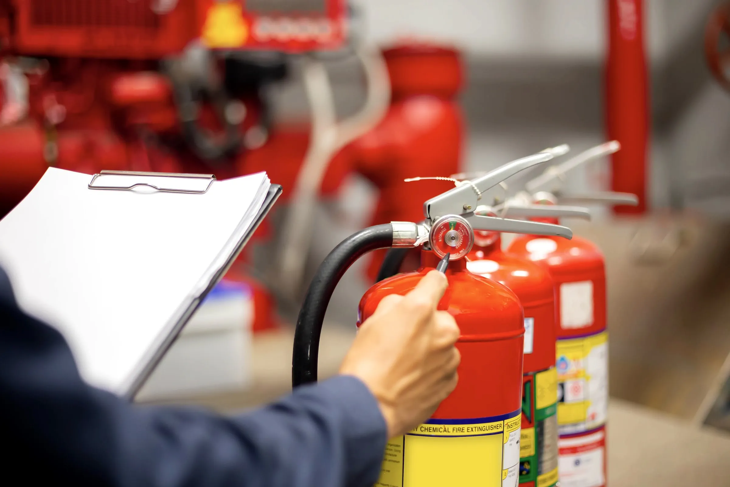 Hand adjusting the pressure gauge on a red fire extinguisher while holding a clipboard, with more extinguishers lined up behind