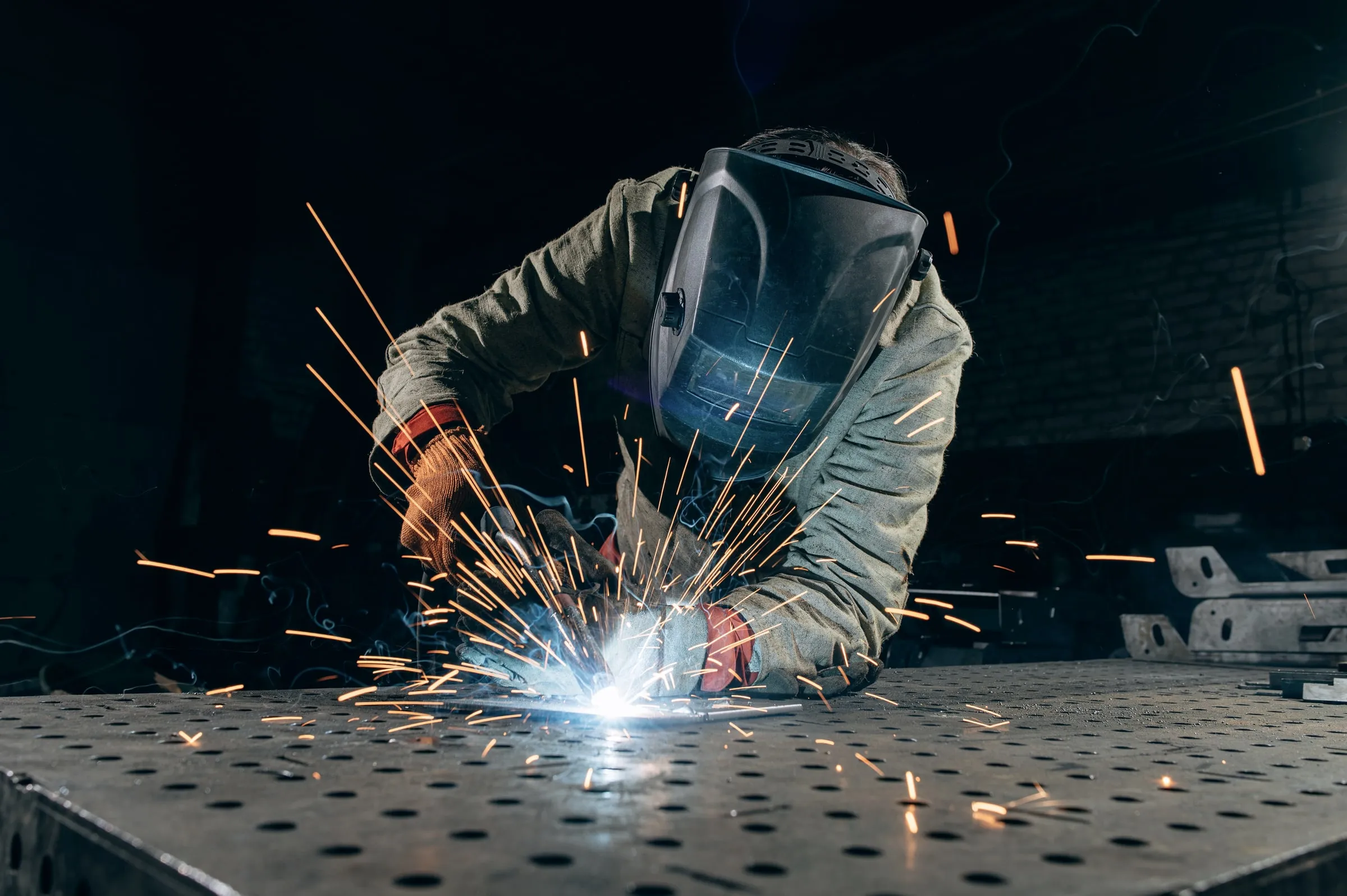 Welder in gray protective gear and a dark visor welding on a perforated steel table, arc and sparks scattering in a workshop