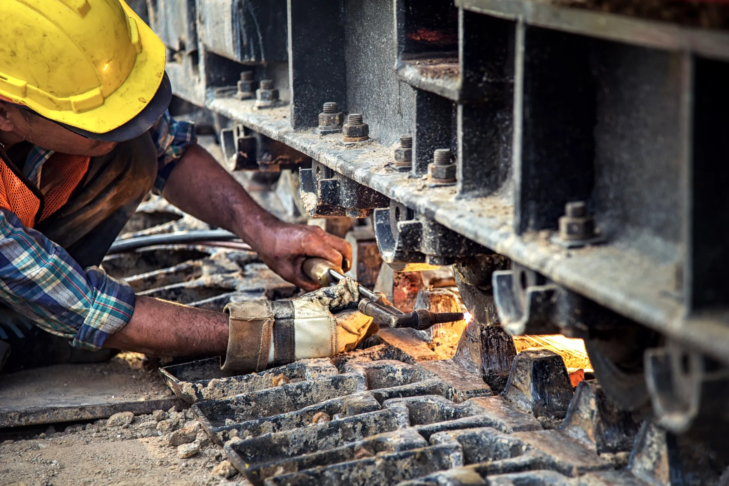 Worker in a yellow hard hat and orange safety vest using a cutting torch on heavy steel machinery, sparks and glowing metal close up