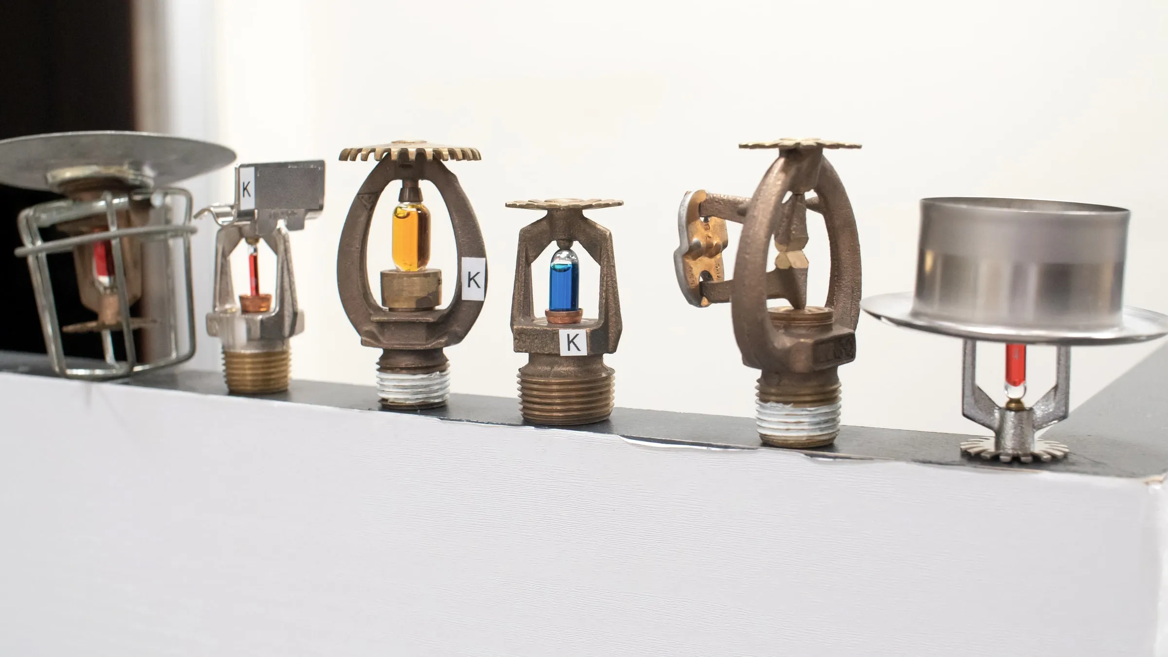 Row of six different fire sprinkler heads of varying styles and finishes displayed side by side on a shelf