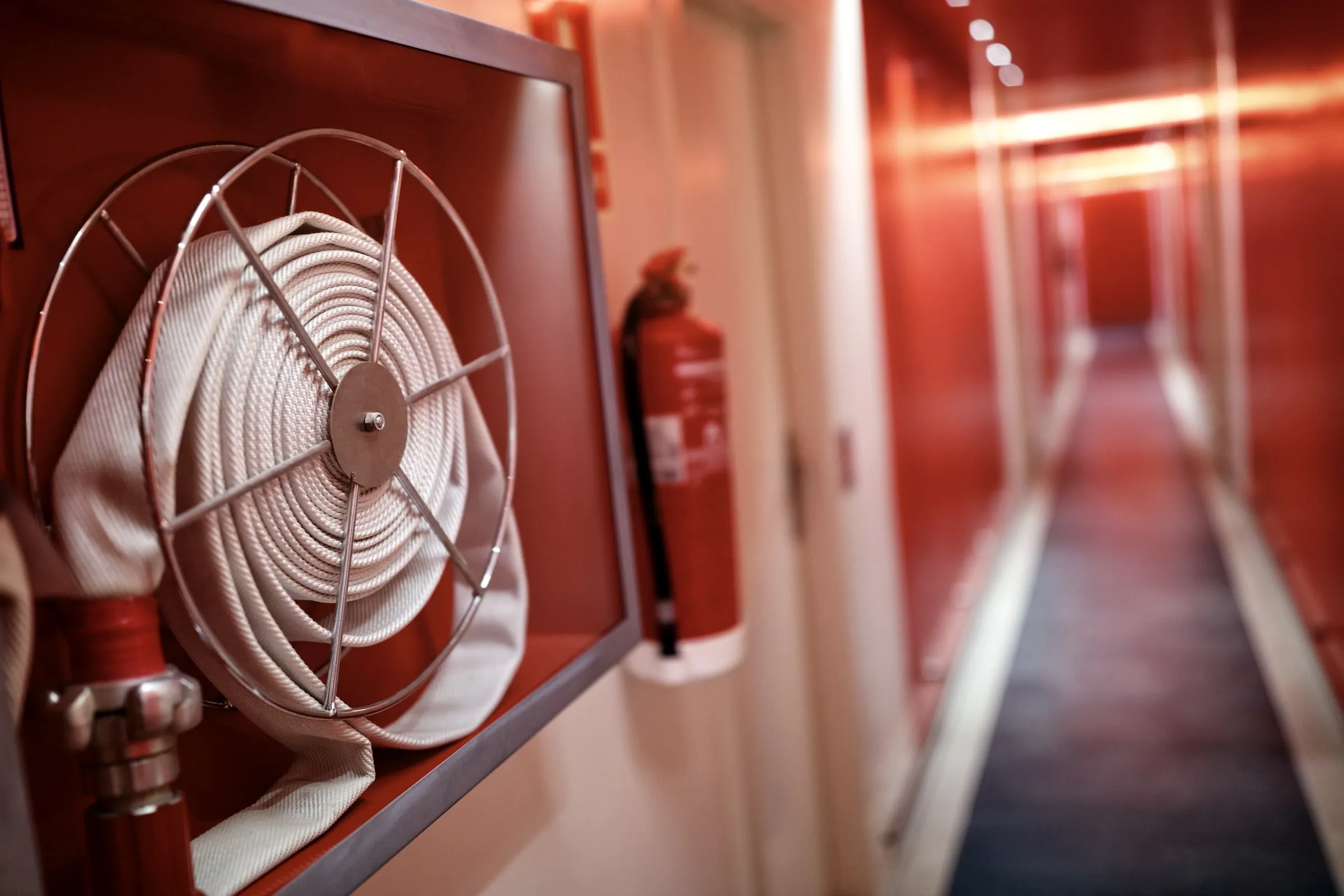 Coiled white fire hose reel and a red fire extinguisher mounted in a wall cabinet along a long red-walled corridor
