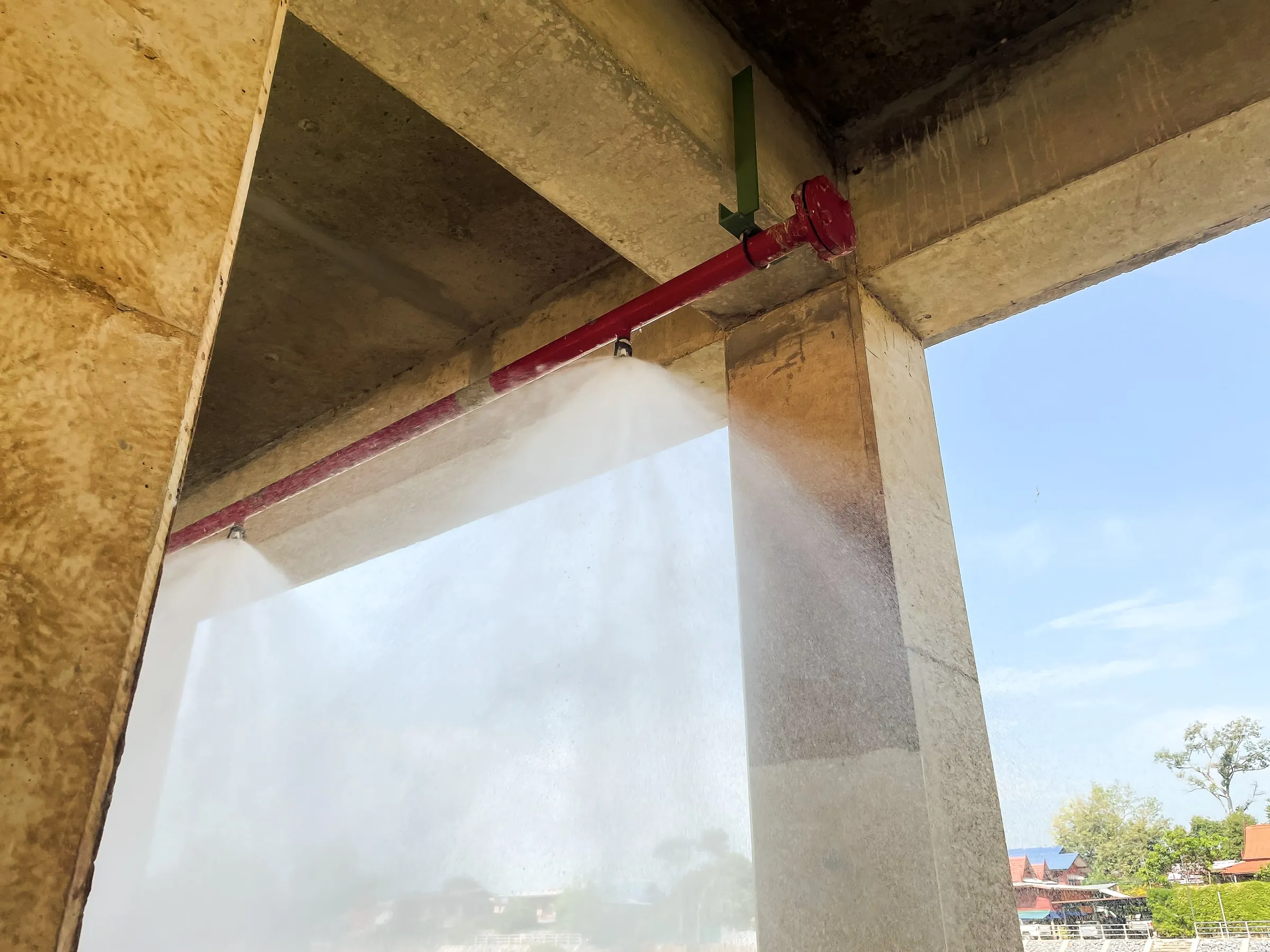 Underside of a concrete parking structure with a red sprinkler pipe actively spraying water during a system test, blue sky beyond