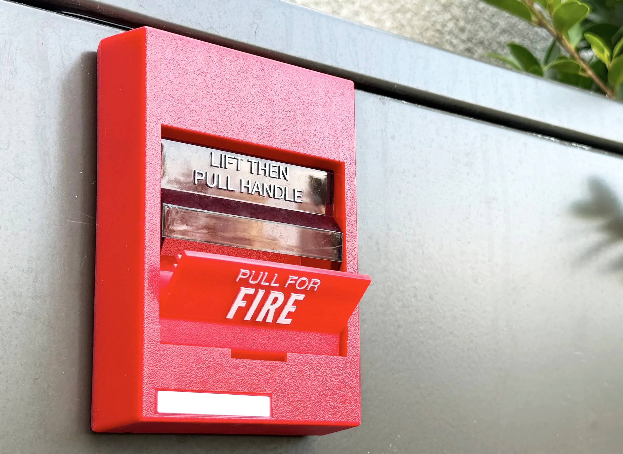 Red fire alarm pull station mounted on an exterior wall reading pull for fire, with greenery visible nearby