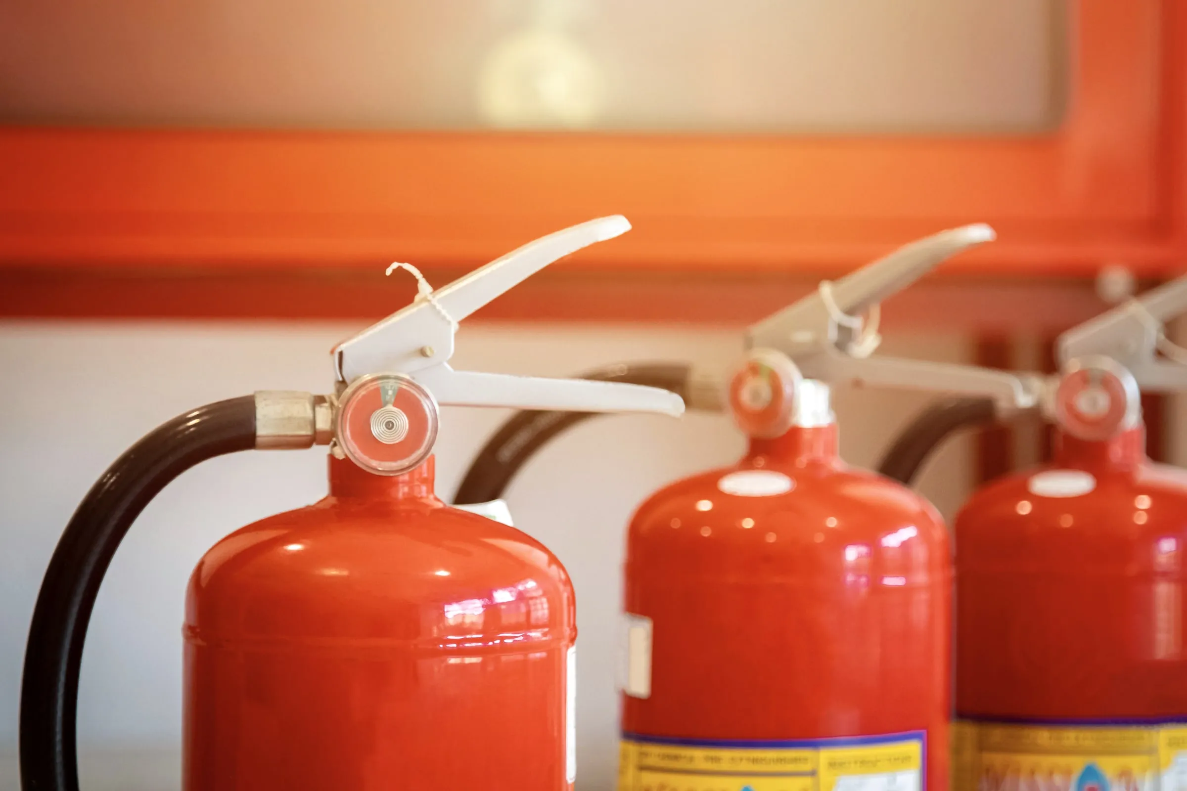 Close-up of three red fire extinguishers with pressure gauges and black hoses mounted in a row against an orange wall