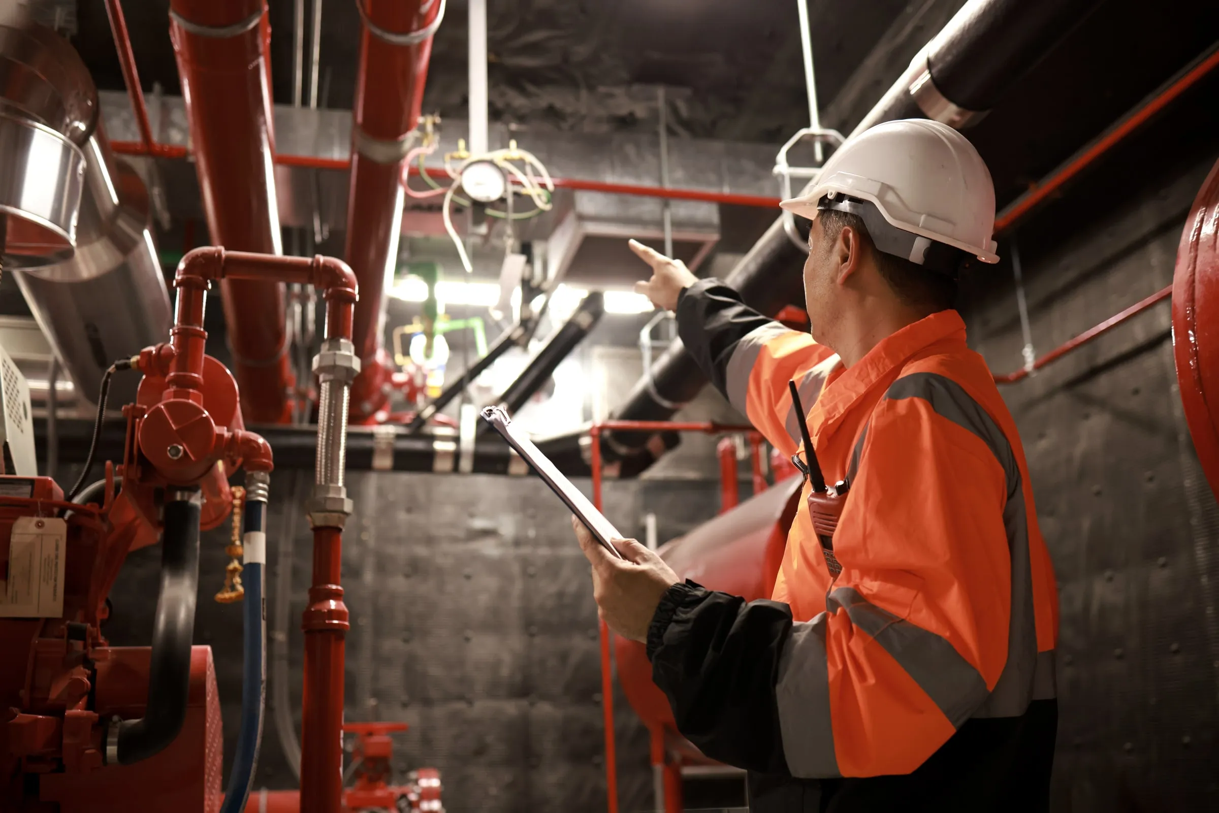 Worker in a hard hat and orange safety jacket points upward while holding a clipboard and radio in a fire pump room