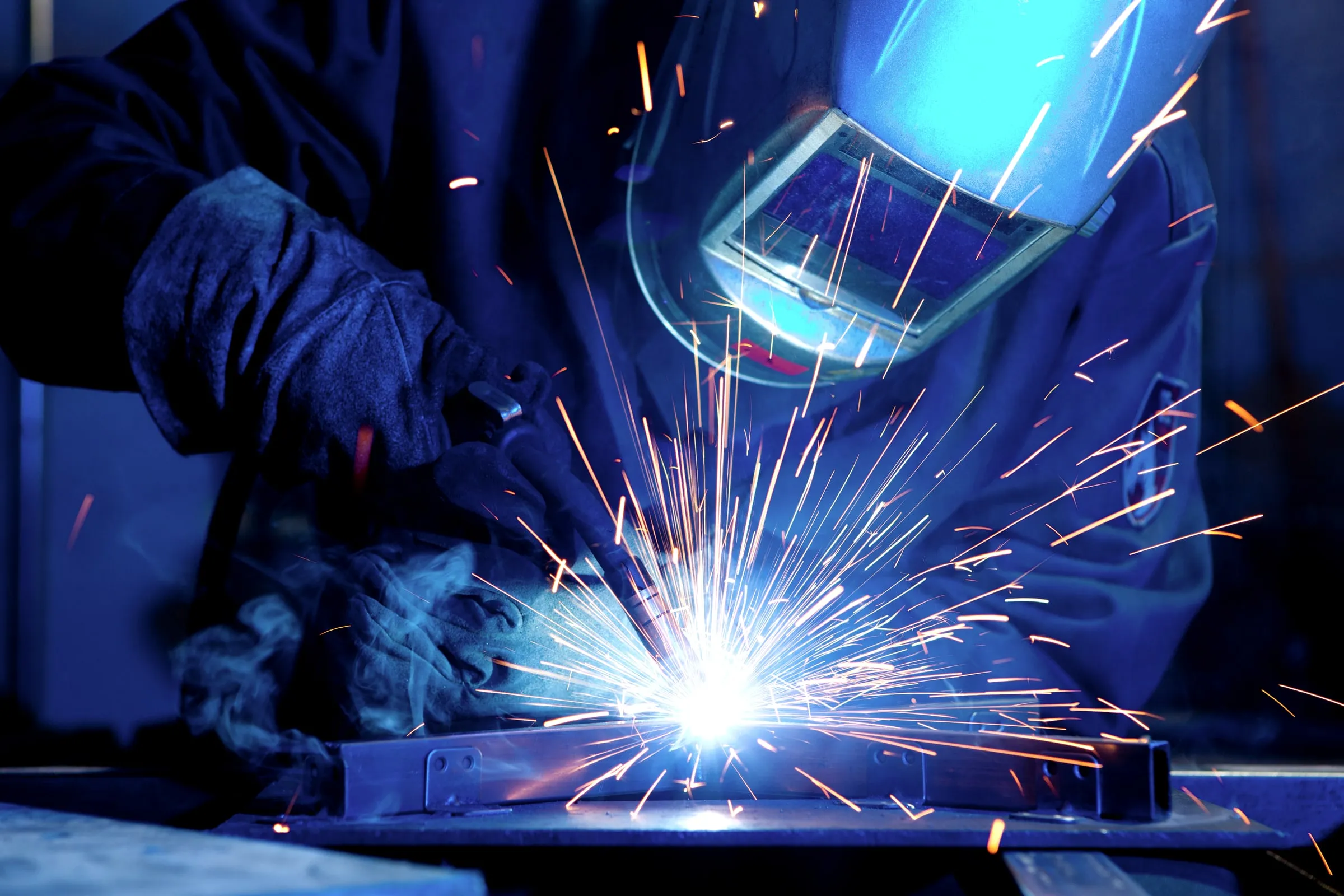 Welder in a dark jacket and blue-tinted helmet running a weld bead on a steel beam, with bright orange sparks scattering