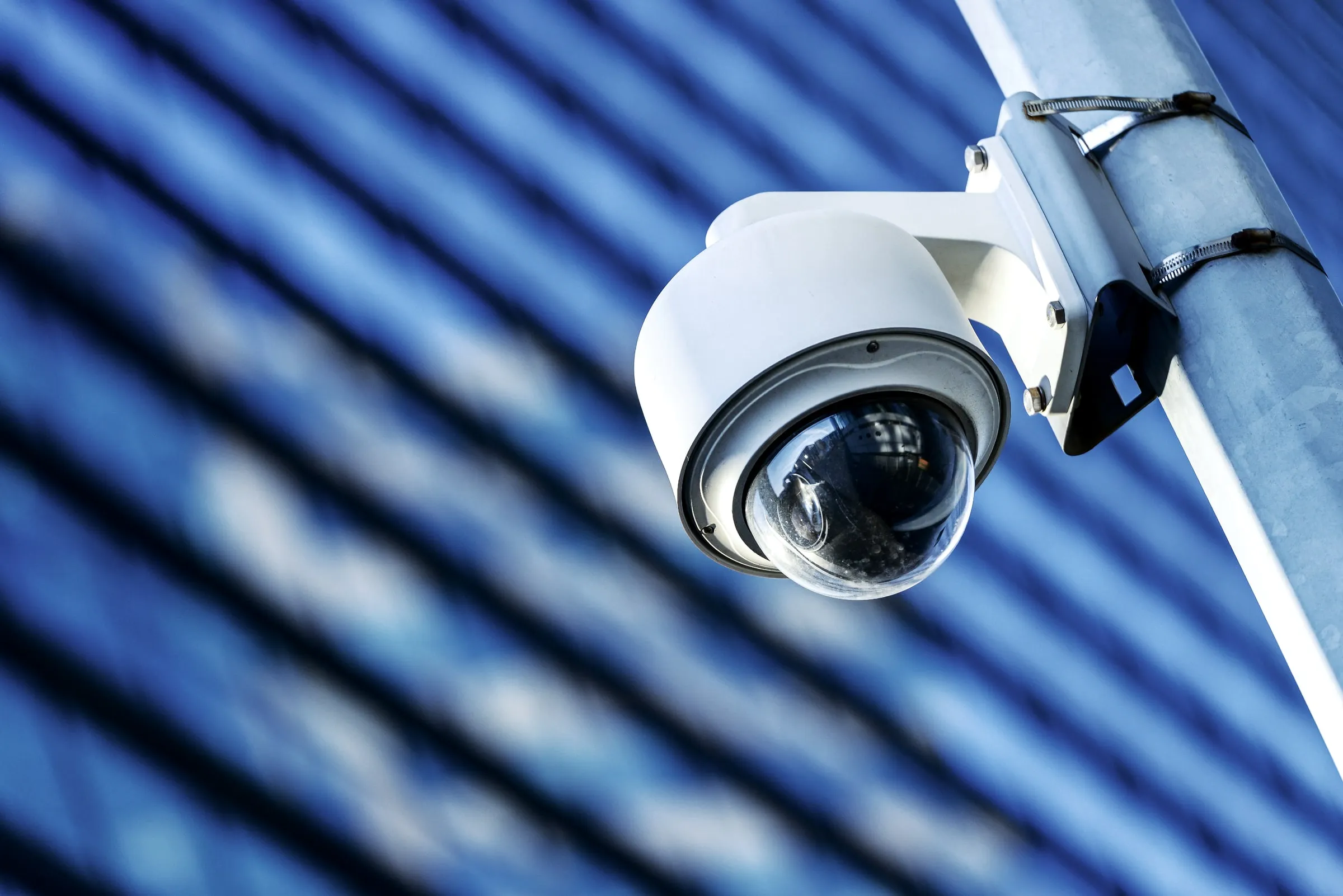 Close view of a white dome security camera mounted on a pole against a blue paneled building facade