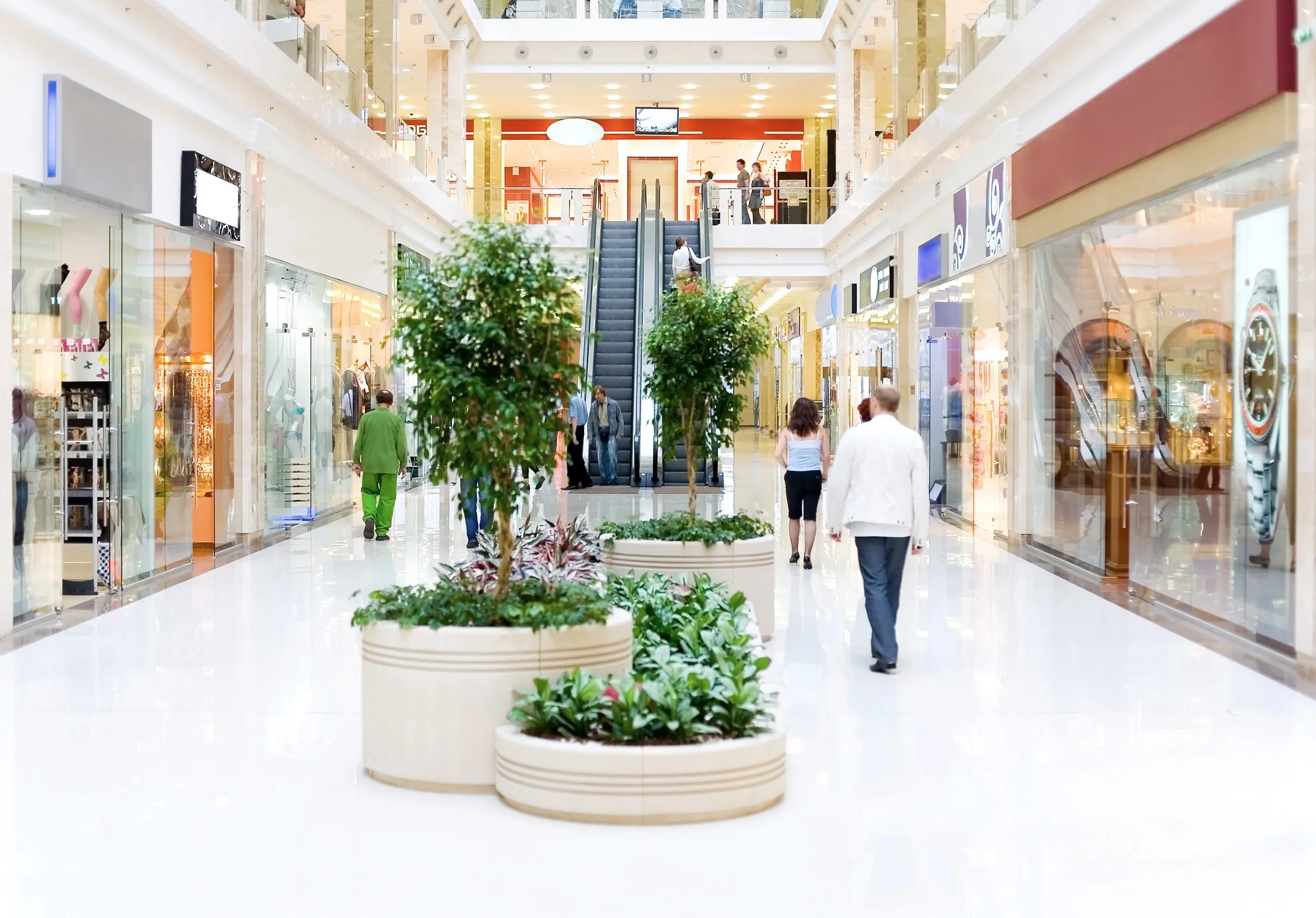 Interior of a shopping mall with escalators, potted plants and shoppers walking past storefronts