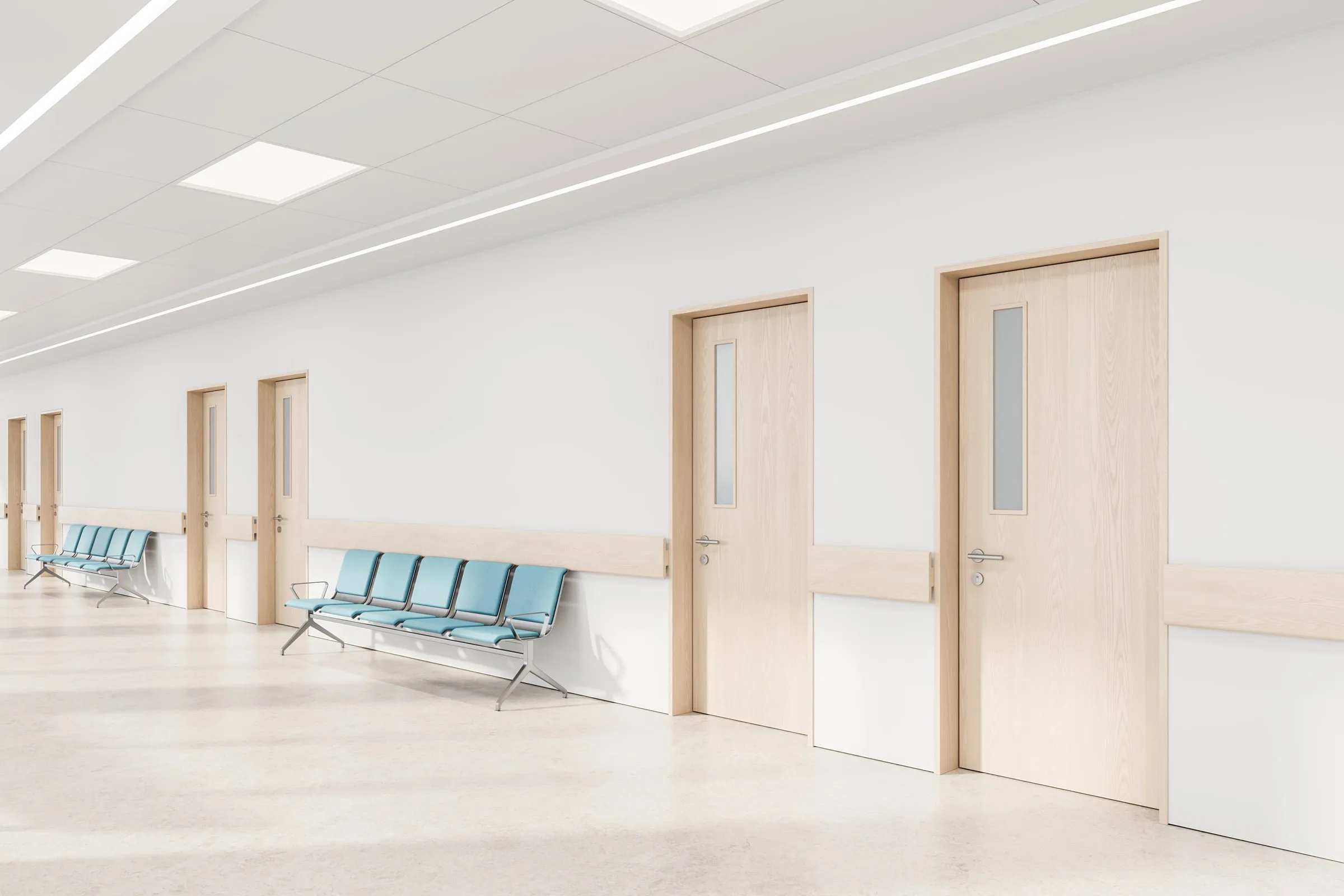 Hospital corridor with wood-toned doors and blue bench seating along a bright, empty hallway