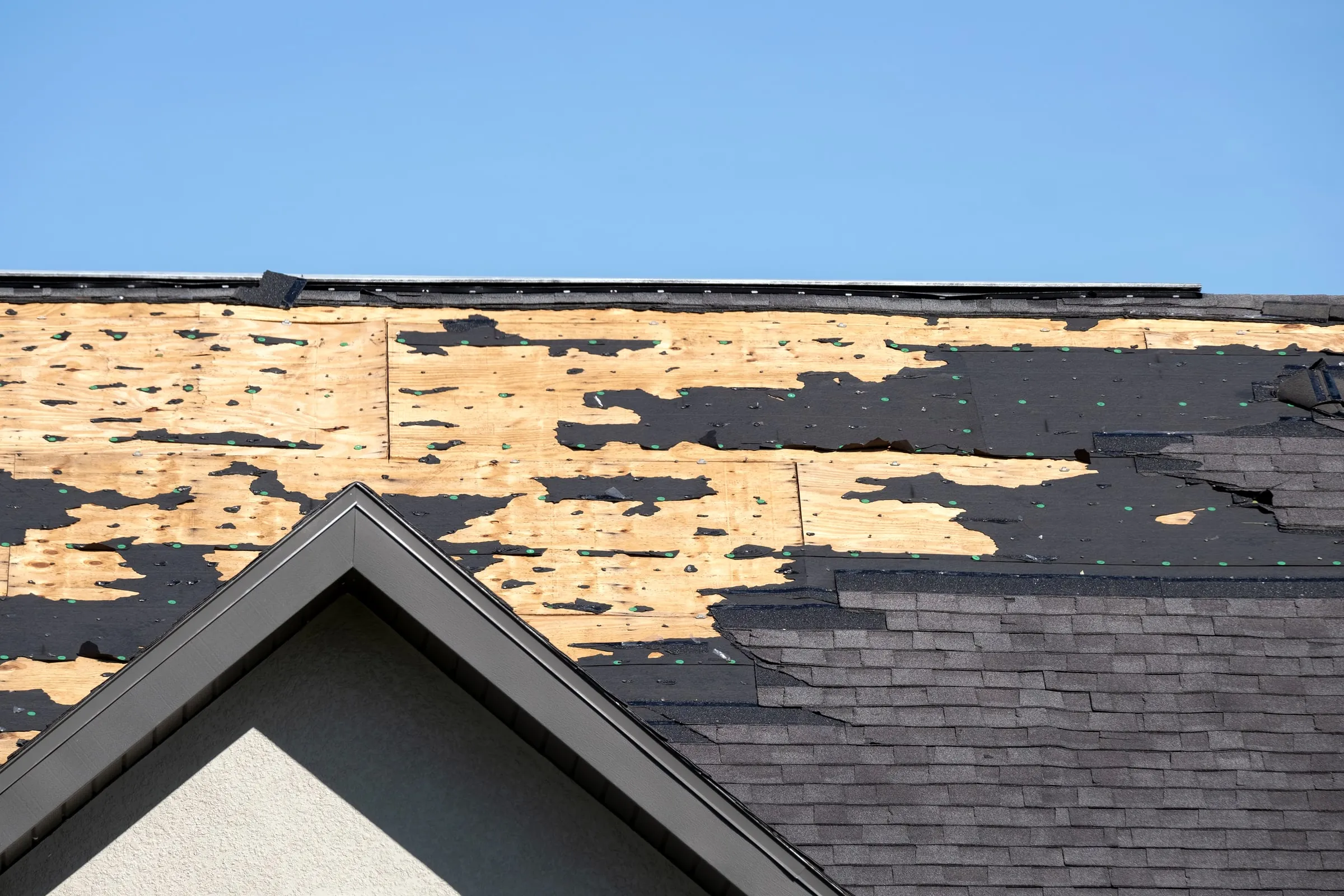 Close-up of a house roof with hurricane wind damage, shingles torn away exposing plywood decking underneath, against a clear blue sky