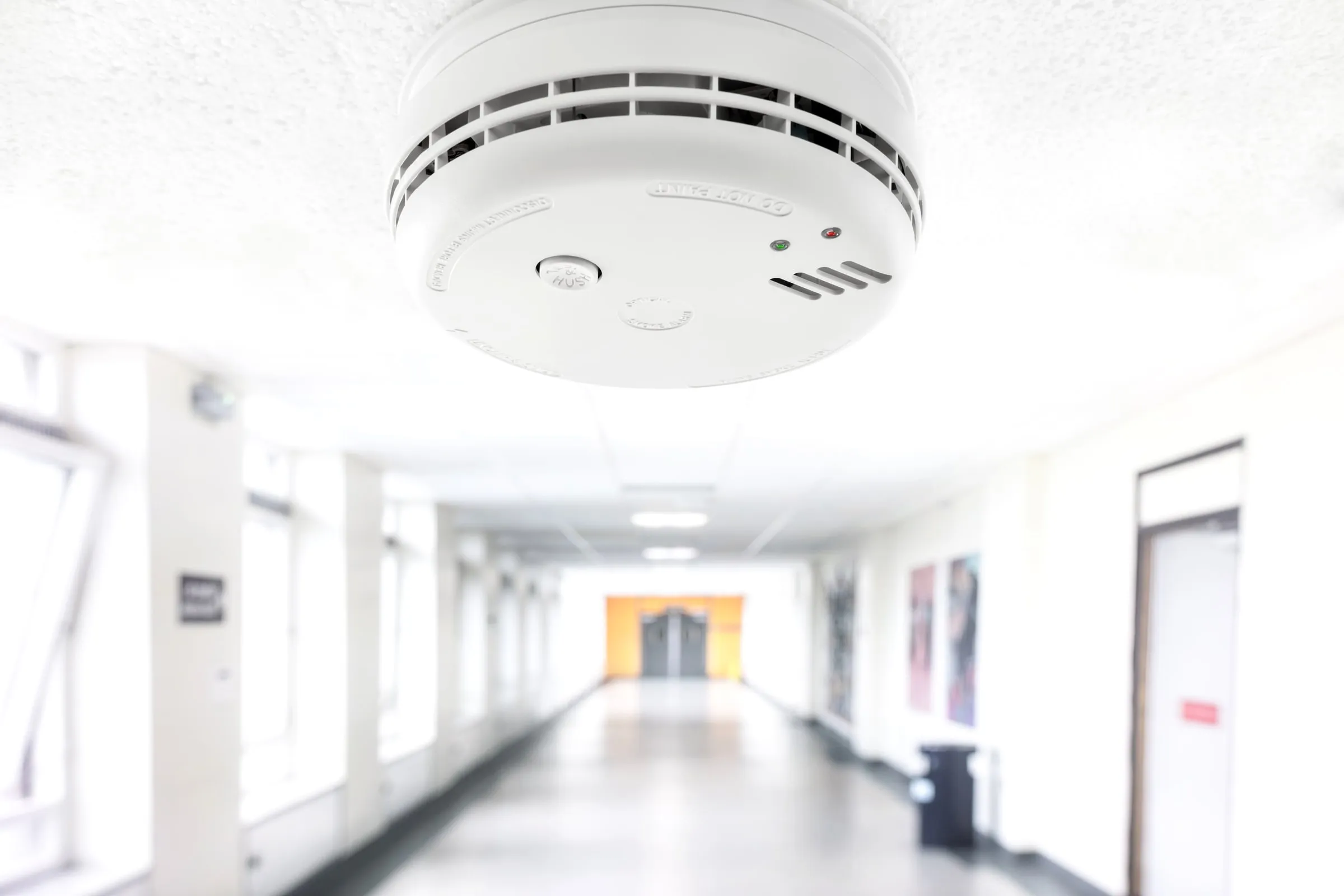White smoke detector mounted on a ceiling above a long, brightly lit institutional corridor with lockers and posters