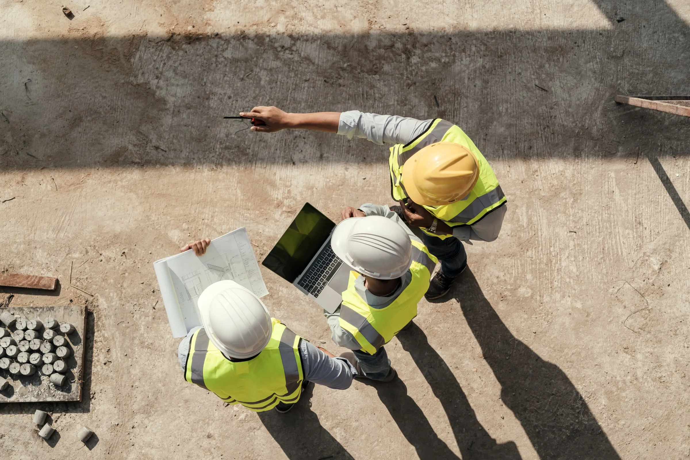 Aerial view of three construction workers in hard hats and safety vests reviewing a blueprint and a laptop on a job site