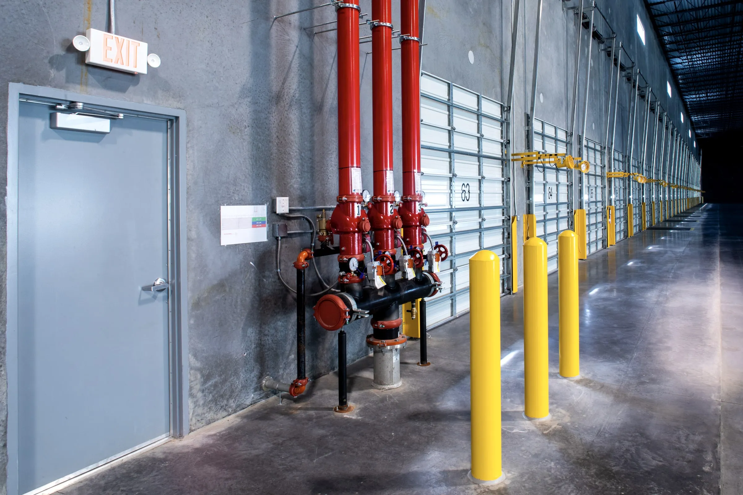 Red sprinkler riser assembly with gauges and valves in a warehouse loading area, yellow bollards and roll-up doors along the wall