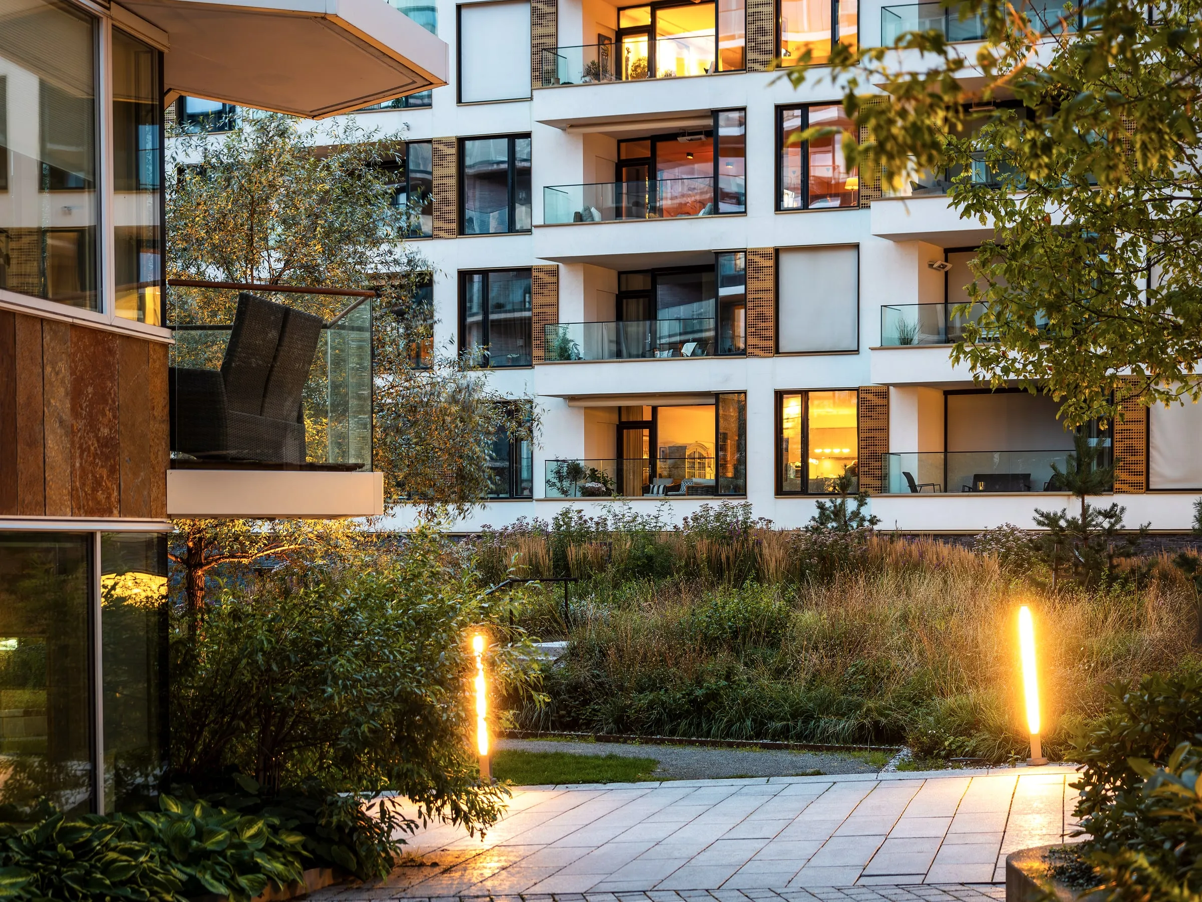 Inner courtyard of a modern apartment building at dusk, with lit balconies, planted beds and bollard lights along a paved path