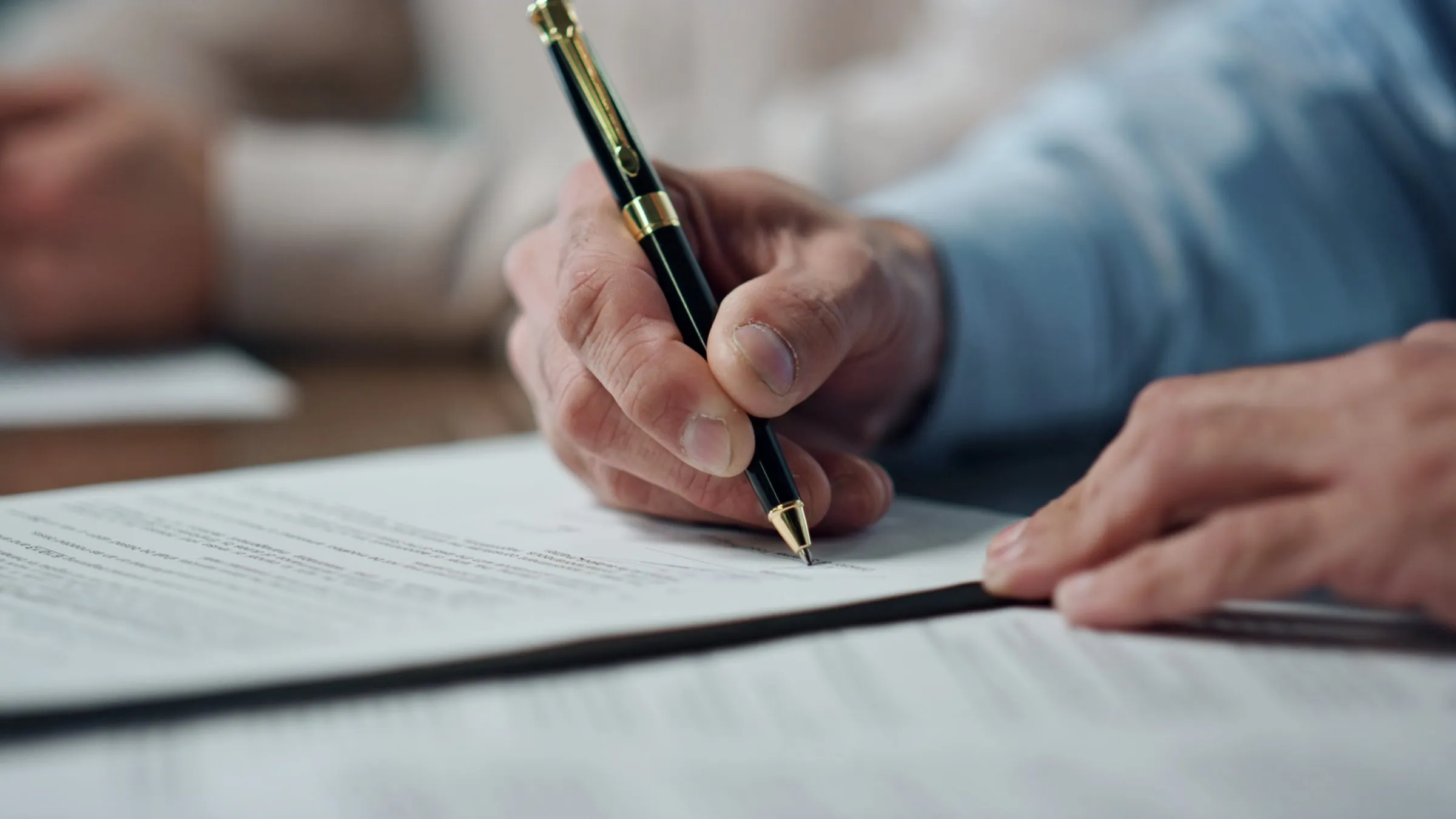 Close-up of a hand signing a printed multi-page document with a fountain pen while a second hand rests on the page nearby
