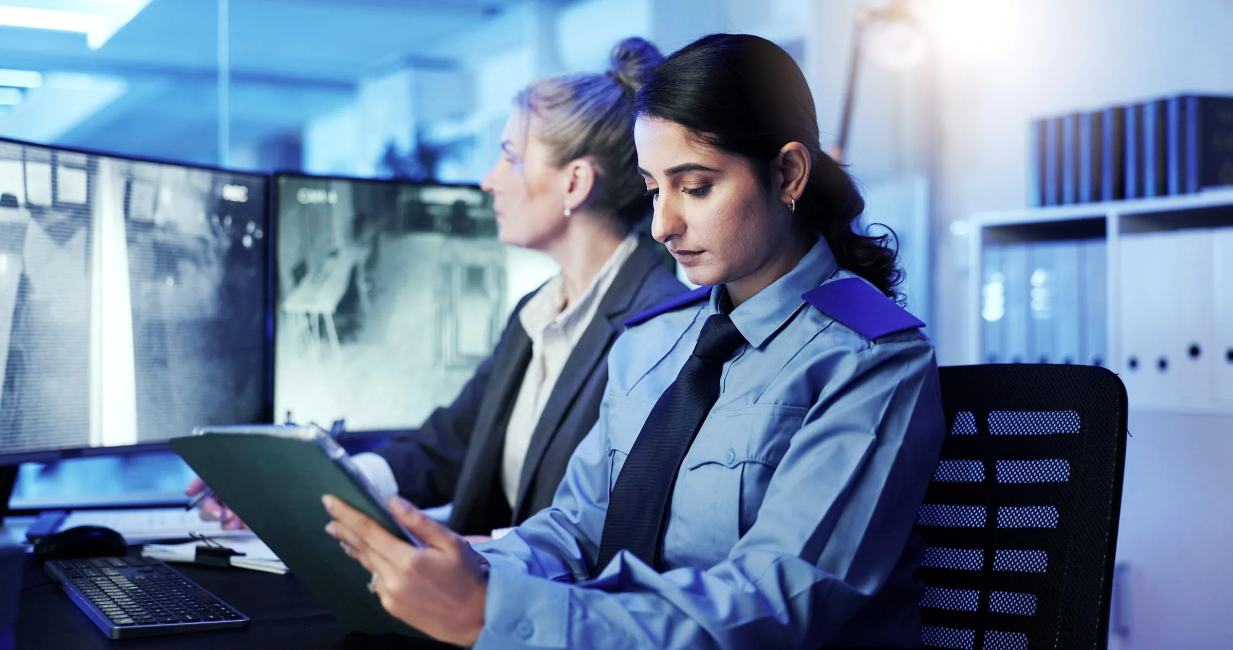 Uniformed security officer reading from a clipboard beside a colleague, with multiple camera monitor screens visible behind them