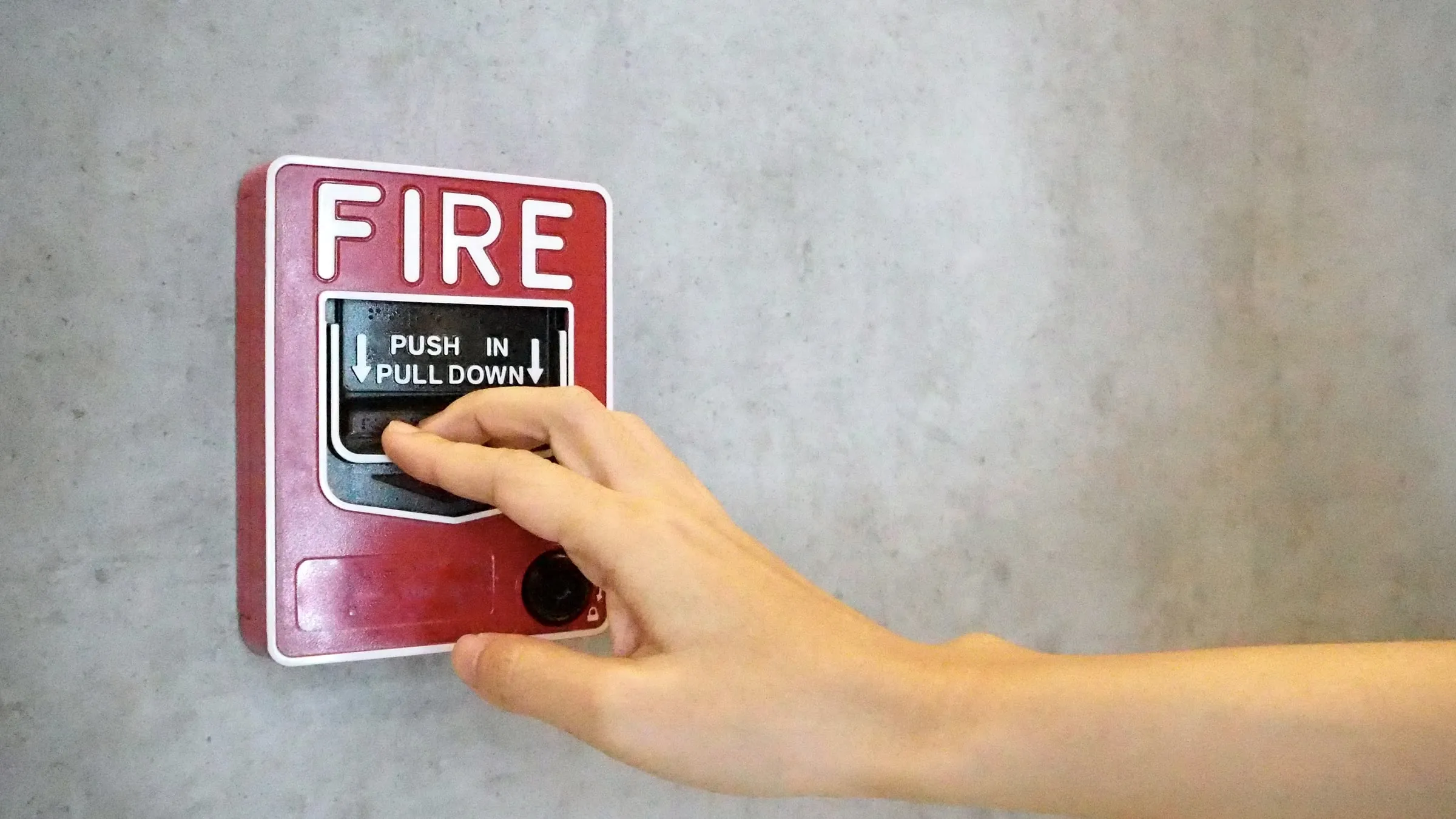 Hand reaching toward a red wall-mounted fire alarm pull station with push-in, pull-down instructions, on a grey concrete wall