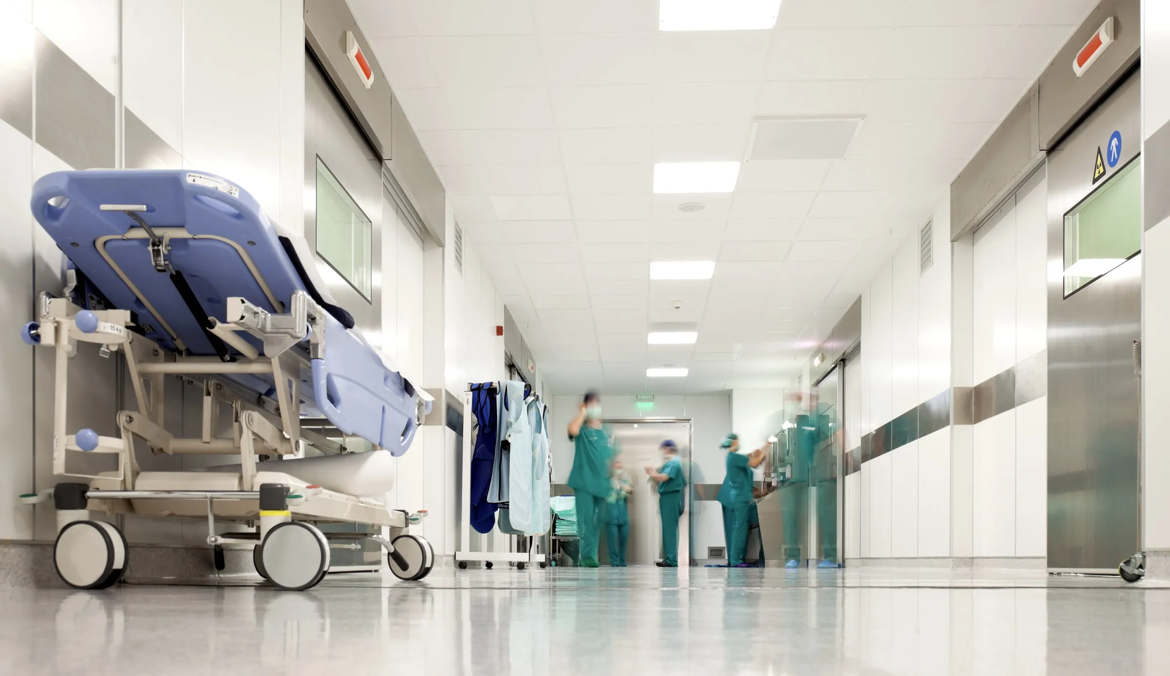 Hospital corridor with an empty gurney in the foreground and medical staff in scrubs standing near a doorway further down the hall