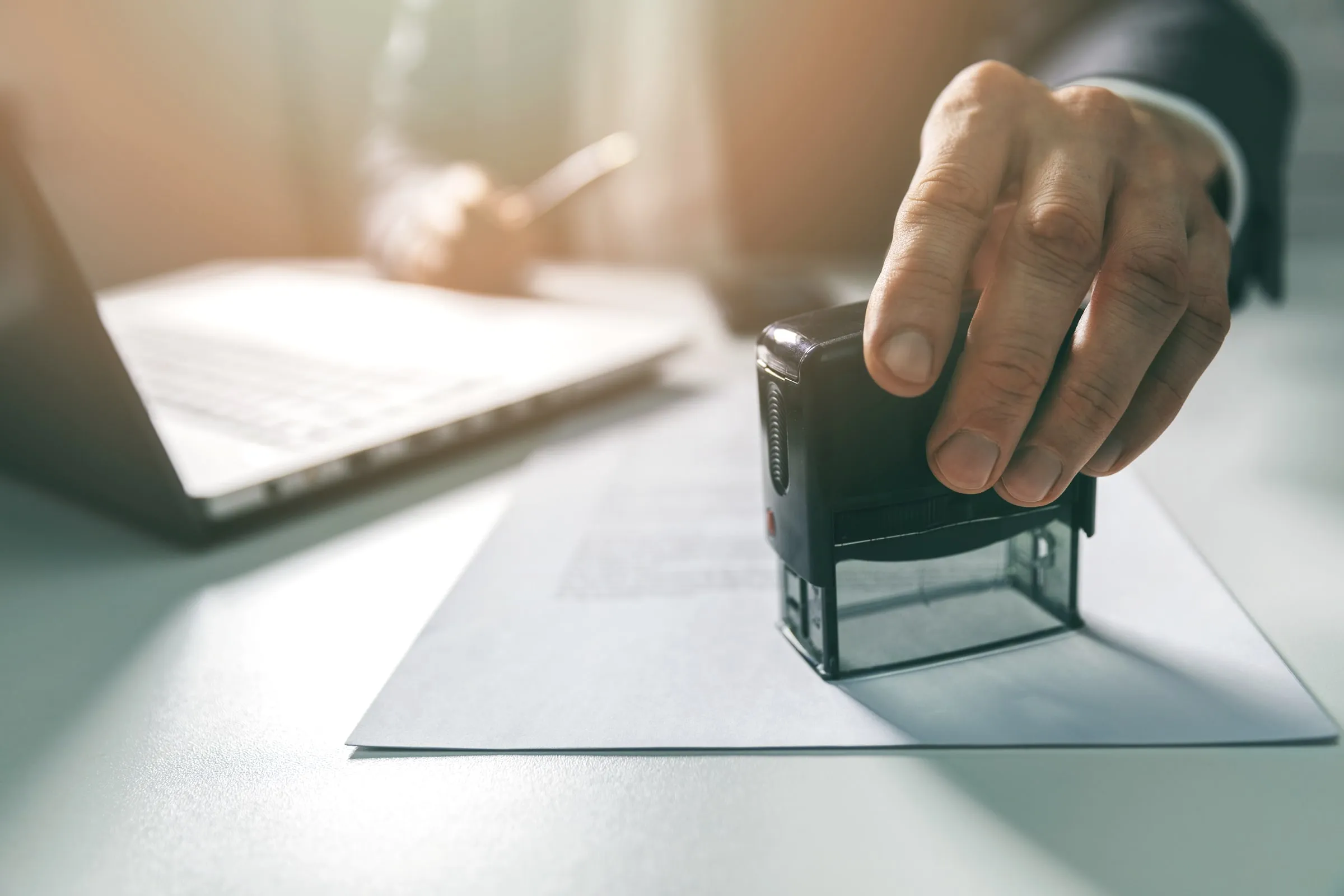 Close view of a hand pressing an official stamp onto a document beside an open laptop in a sunlit office