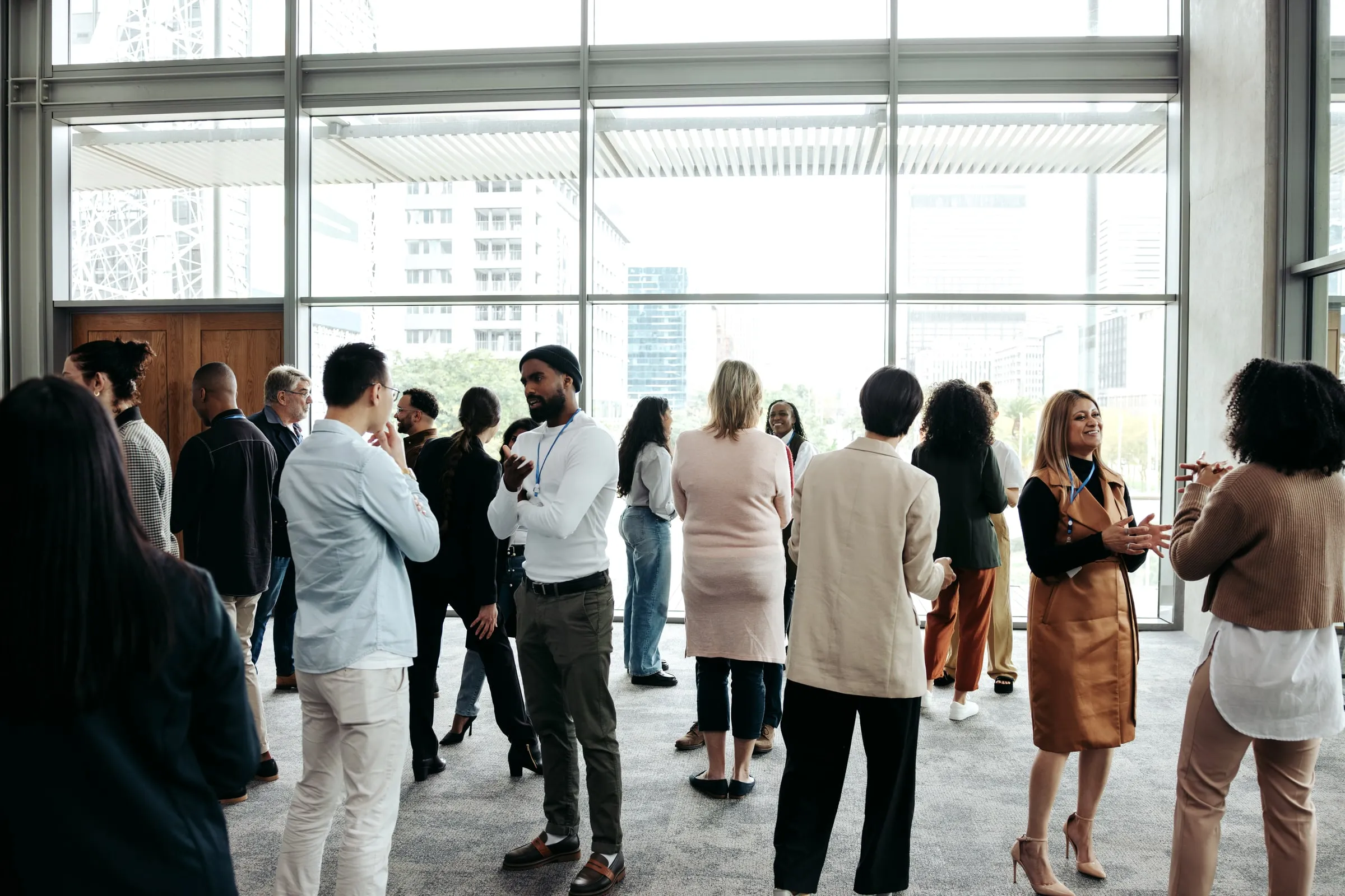 Group of businesspeople with lanyards mingling in a glass-walled lobby with a city skyline visible outside