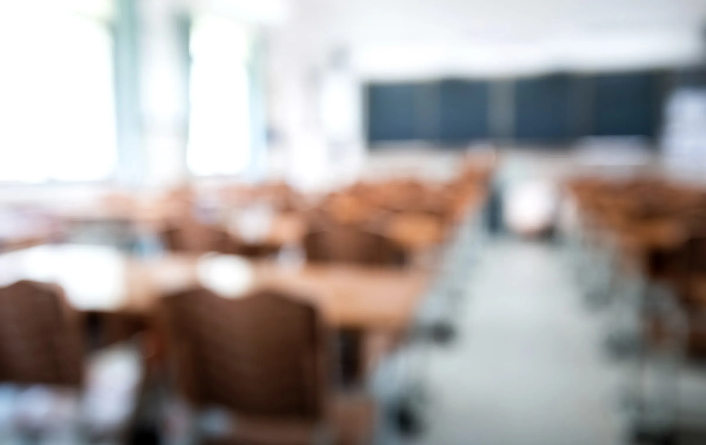 Empty classroom shown in soft focus, rows of desks and chairs facing a dark board beneath bright windows